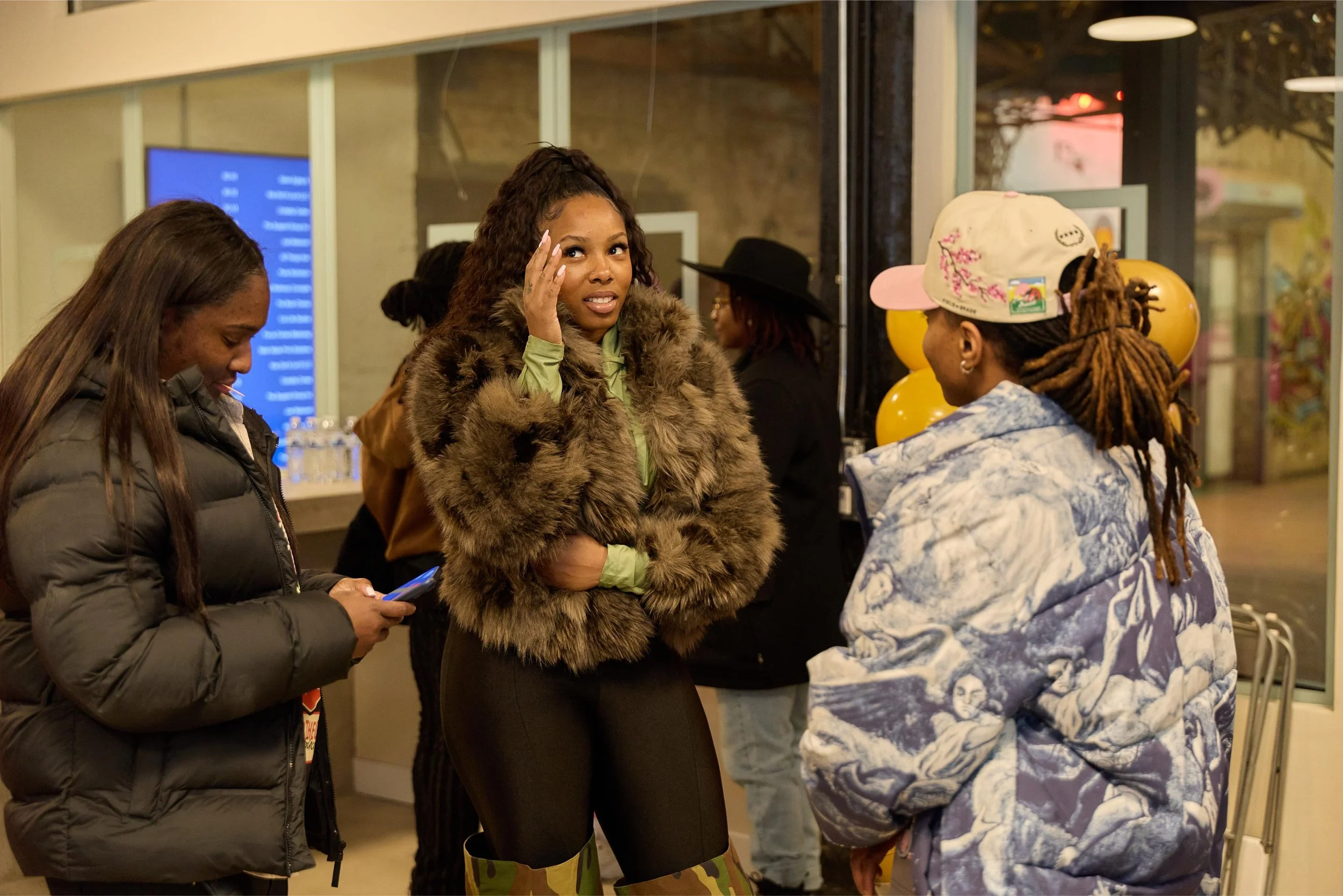 Group of women talking inside a cafe or restaurant, with some wearing jackets and one in a faux fur coat. One woman looks surprised or engaged, while another is focused on her phone. The background has balloons and vibrant decor at Jem Junkies Tooth 