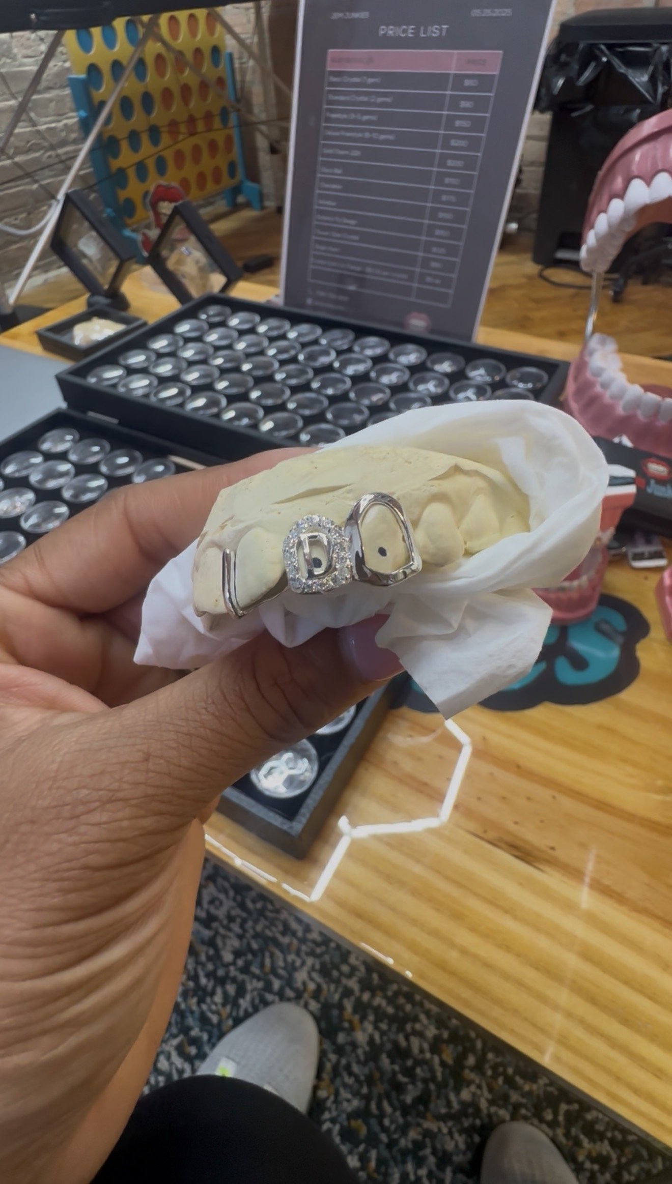 Close-up of a person holding a dental model with a decorative 'LOVE' ring, in a setting with a price list, connect four game, and decorative items on a wooden table.