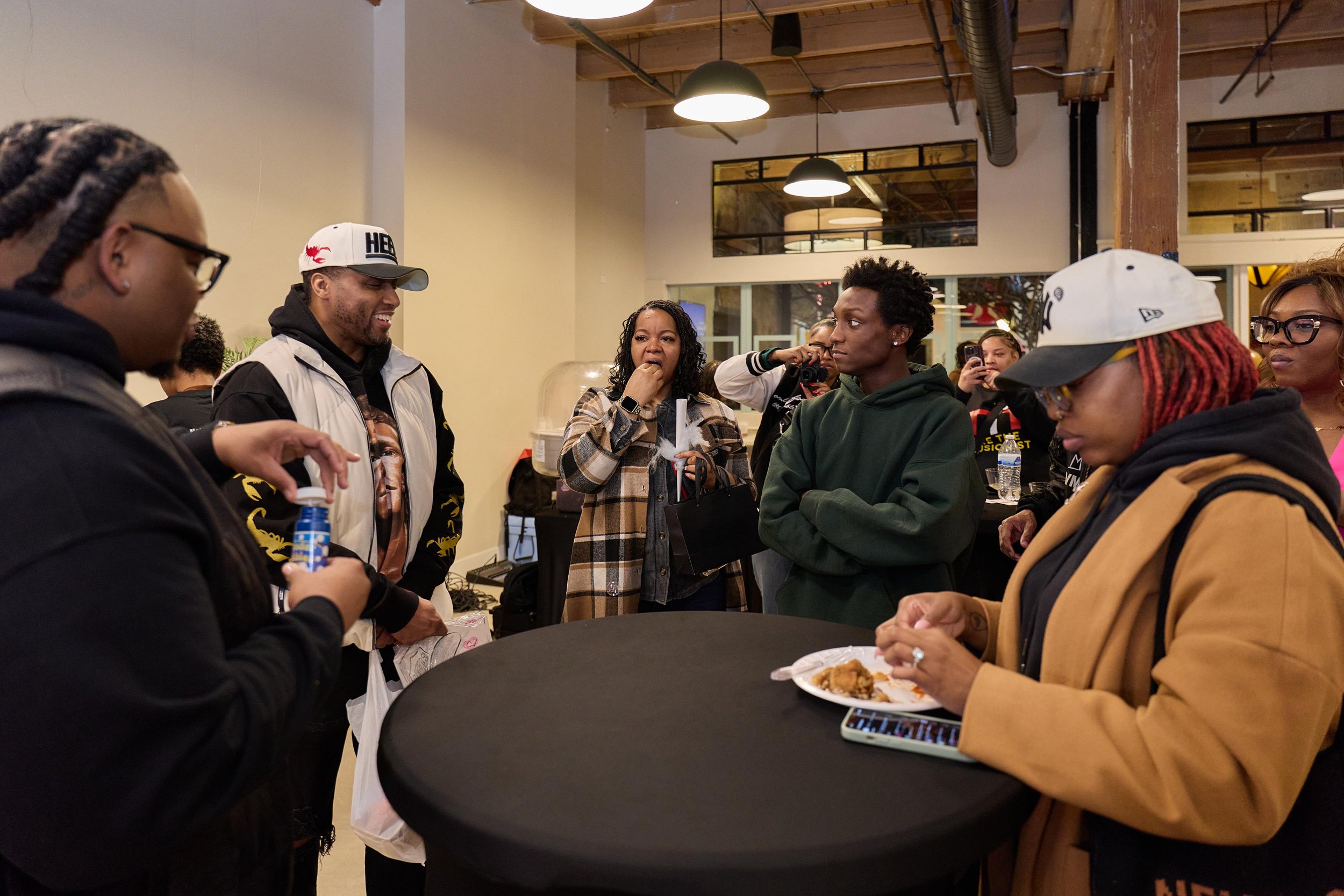 Group of people socializing at a casual indoor event around a black table, some eating, some taking photos, with warm lighting and exposed ceiling beams at Jem Junkies Tooth Gems and  Custom Grillz in Chicago 