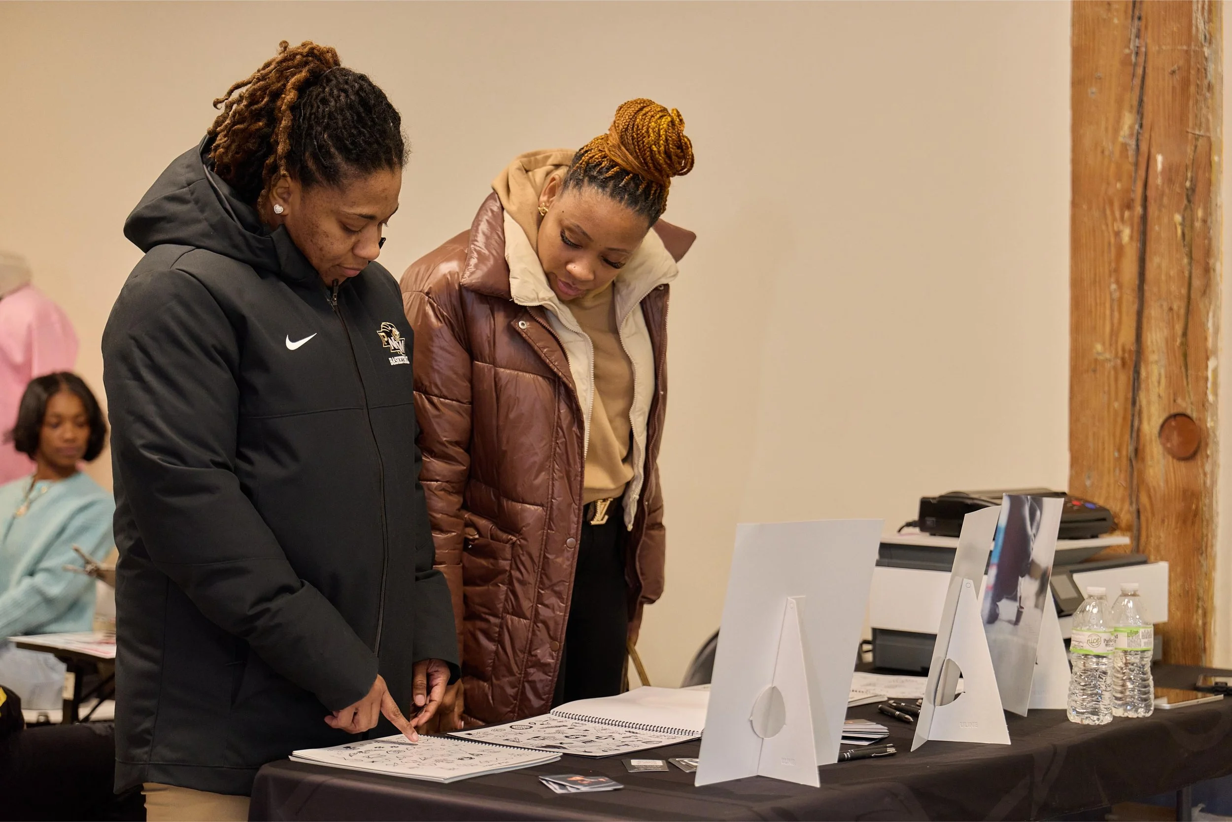 Two women are looking at printed materials on a table, with one pointing at the papers. There are three water bottles and three white display boards on the table, and other people can be seen in the background.