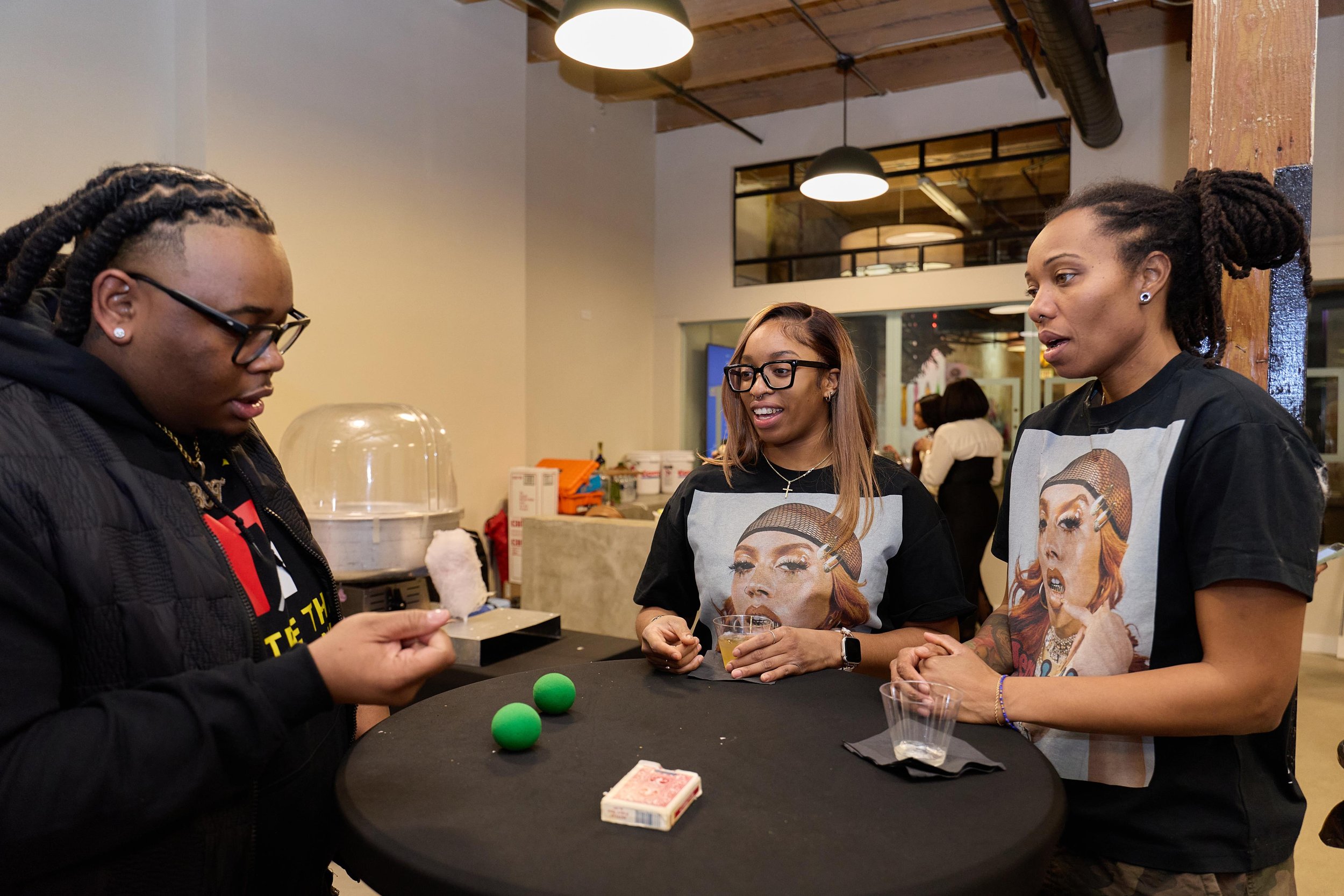 Three women gathered around a black table in a casual indoor setting, with one woman holding cotton candy and the other two watching and smiling at Jem Junkies Tooth Gems and  Custom Grillz in Chicago 