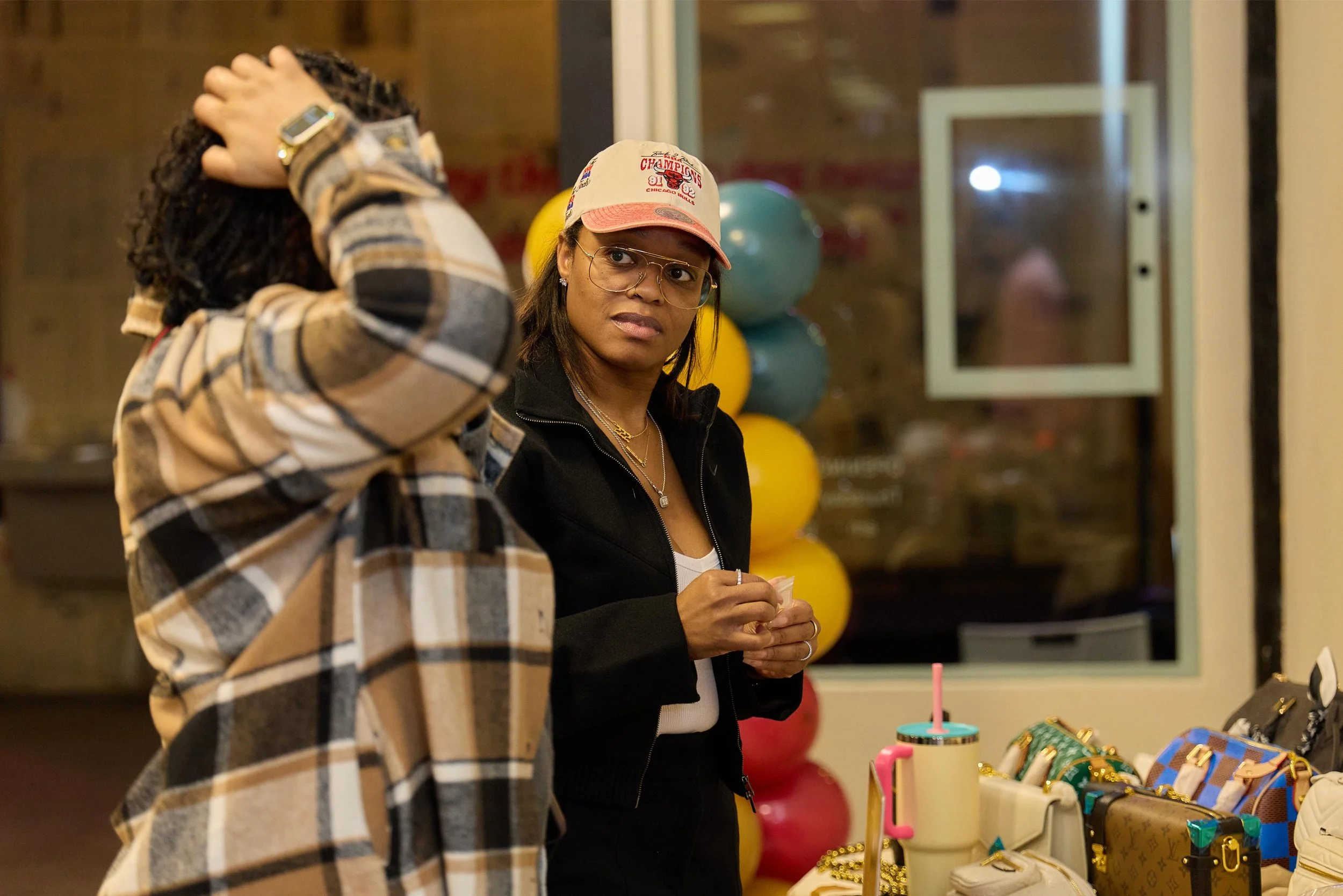 Two women are conversing at a store display with handbags and accessories. One woman wears a plaid jacket, and the other wears glasses, a pink cap, and a black jacket. Balloons are visible in the background.