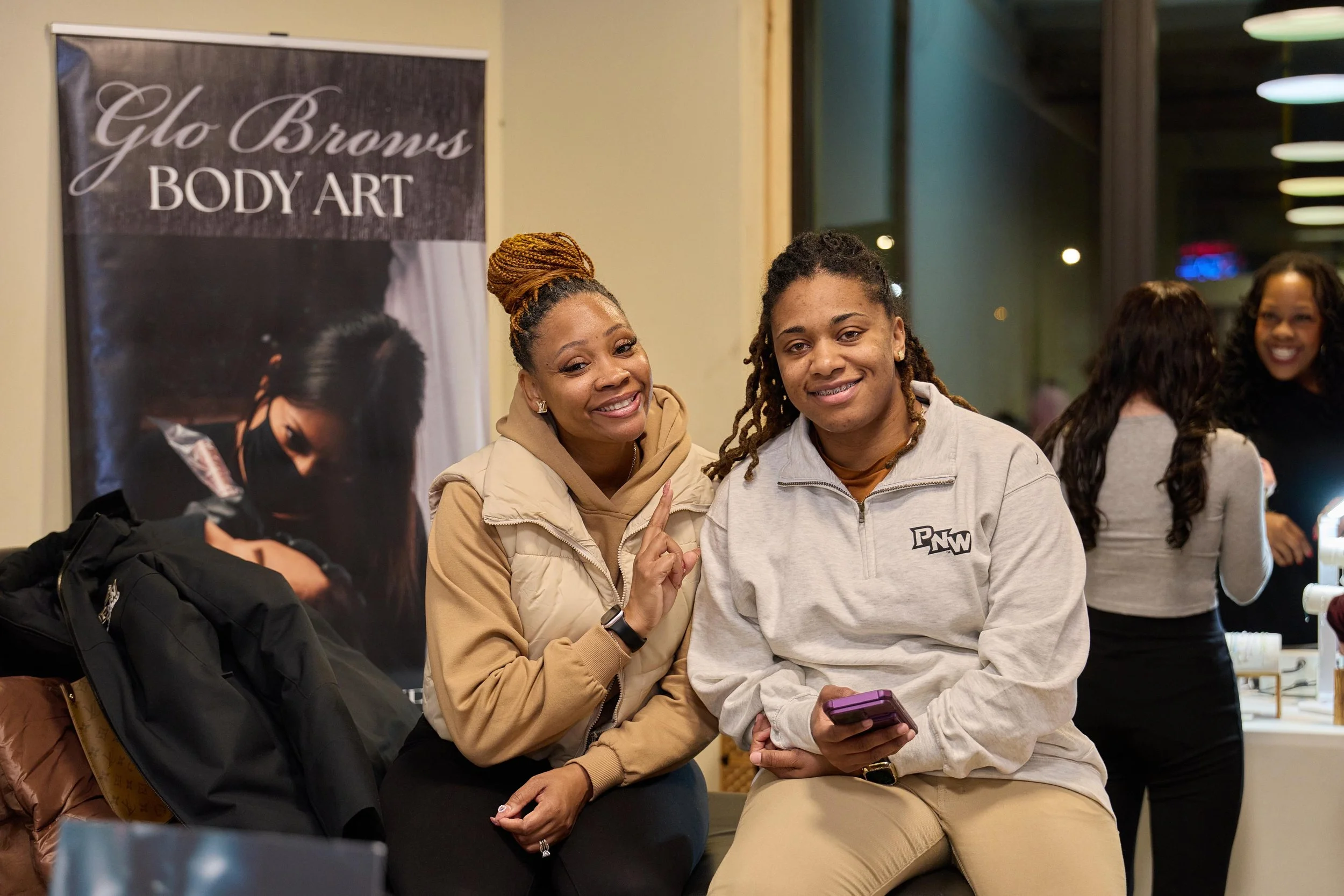 Two women sitting together at a makeup and body art event, smiling at the camera. A sign behind them reads 'Glo Browns Body Art'. at Jem Junkies Tooth Gems and  Custom Grillz in Chicago
