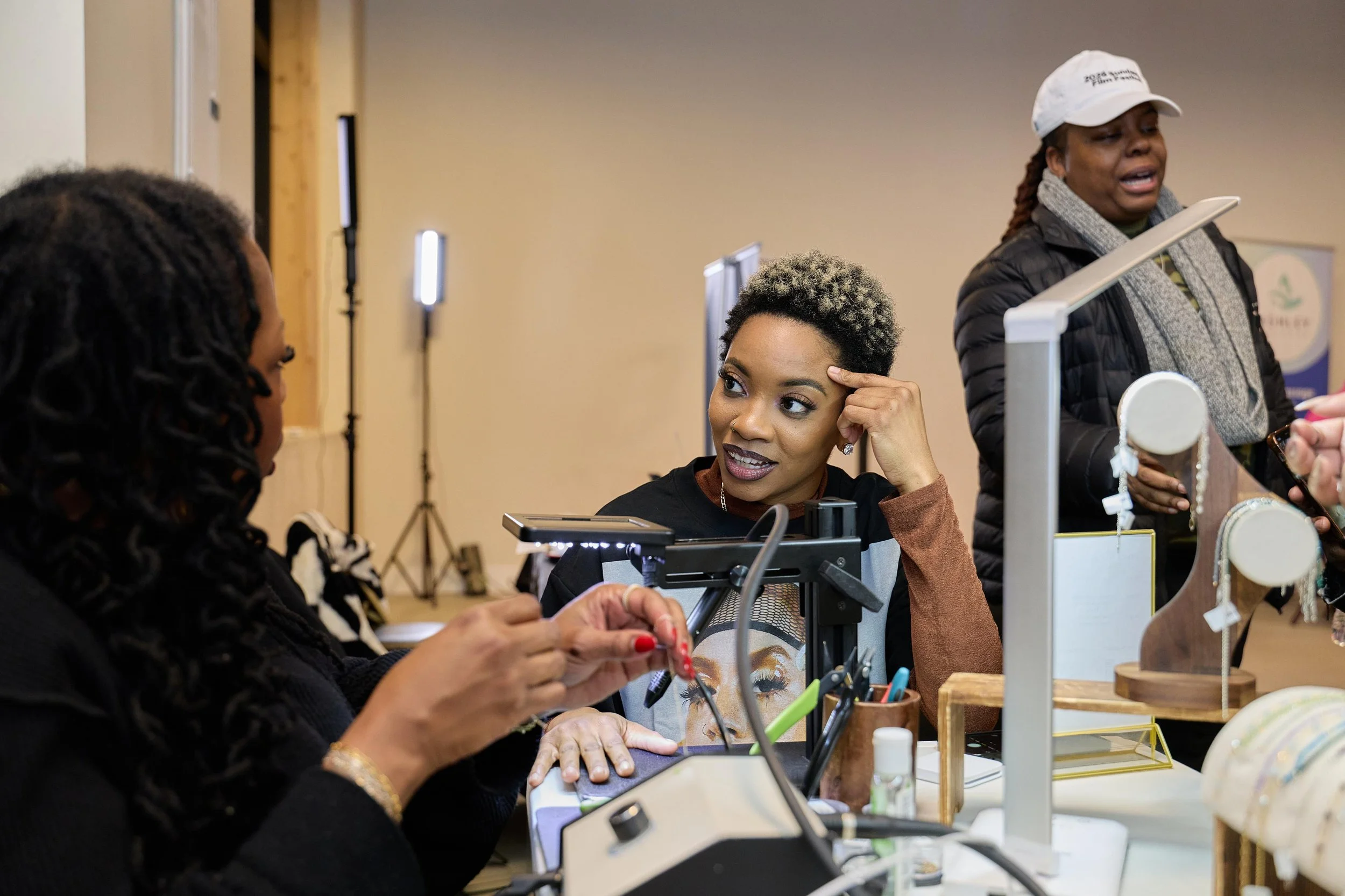Three women engaging in a conversation at a craft fair or jewelry booth. One woman is showing jewelry while another woman is listening and talking, and the third woman is walking past the booth.