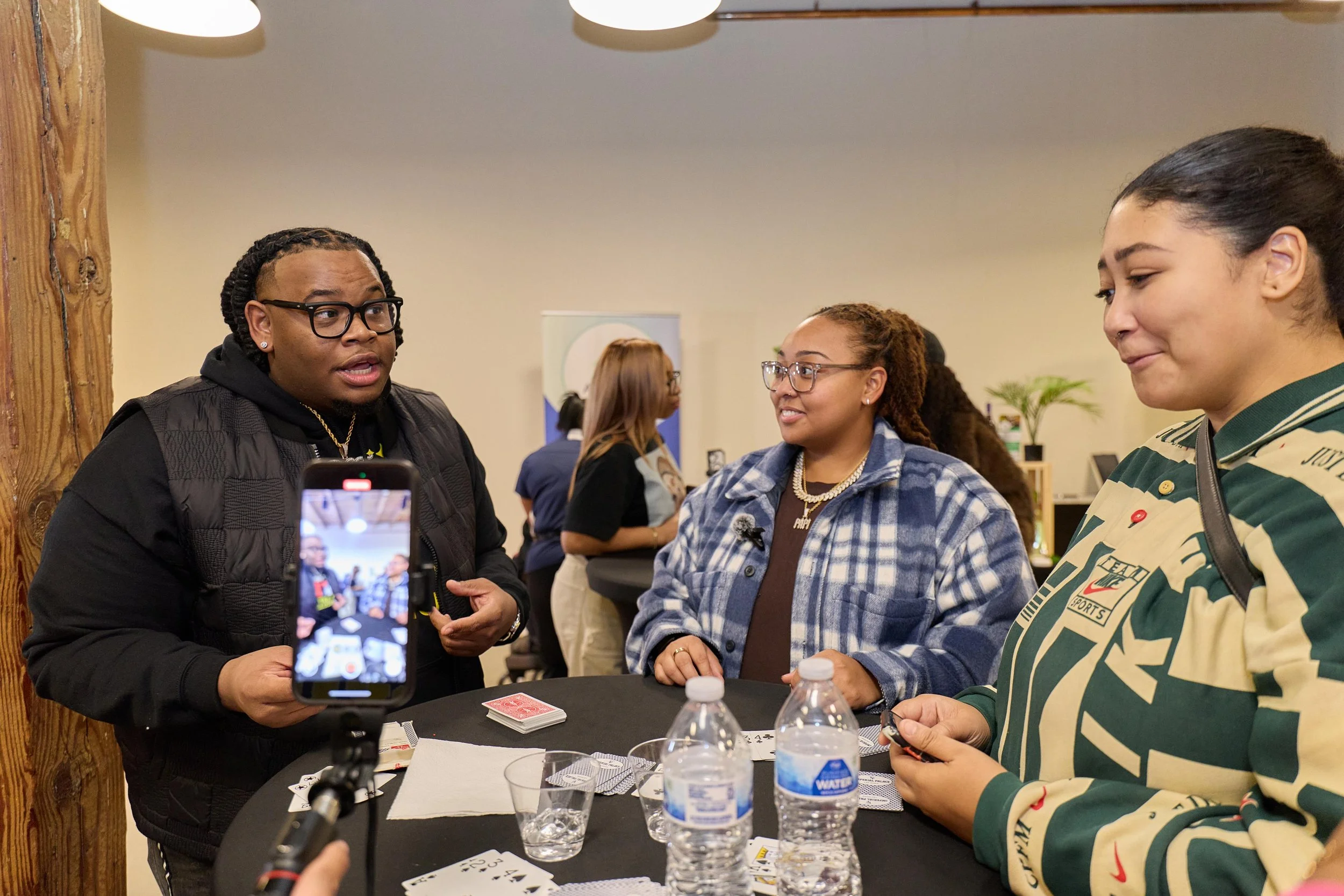 Three women standing around a table with playing cards and water bottles, with a person recording the scene with a smartphone. The woman on the left is speaking, the woman in the middle is smiling, and the woman on the right is holding cards.