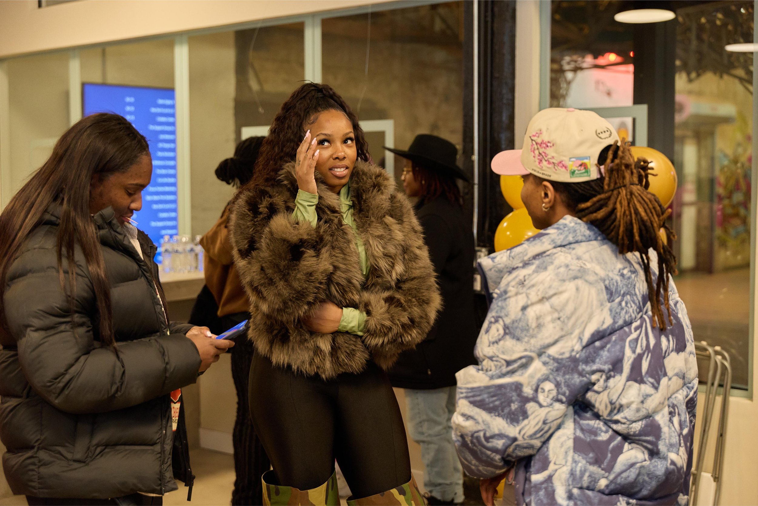 Group of women socializing indoors, one woman with long braids, wearing a white baseball cap with patches, a blue patterned jacket, and camouflage boots talks to a woman with curly hair in a fur jacket at Jem Junkies Tooth Gems and  Custom Grillz in 