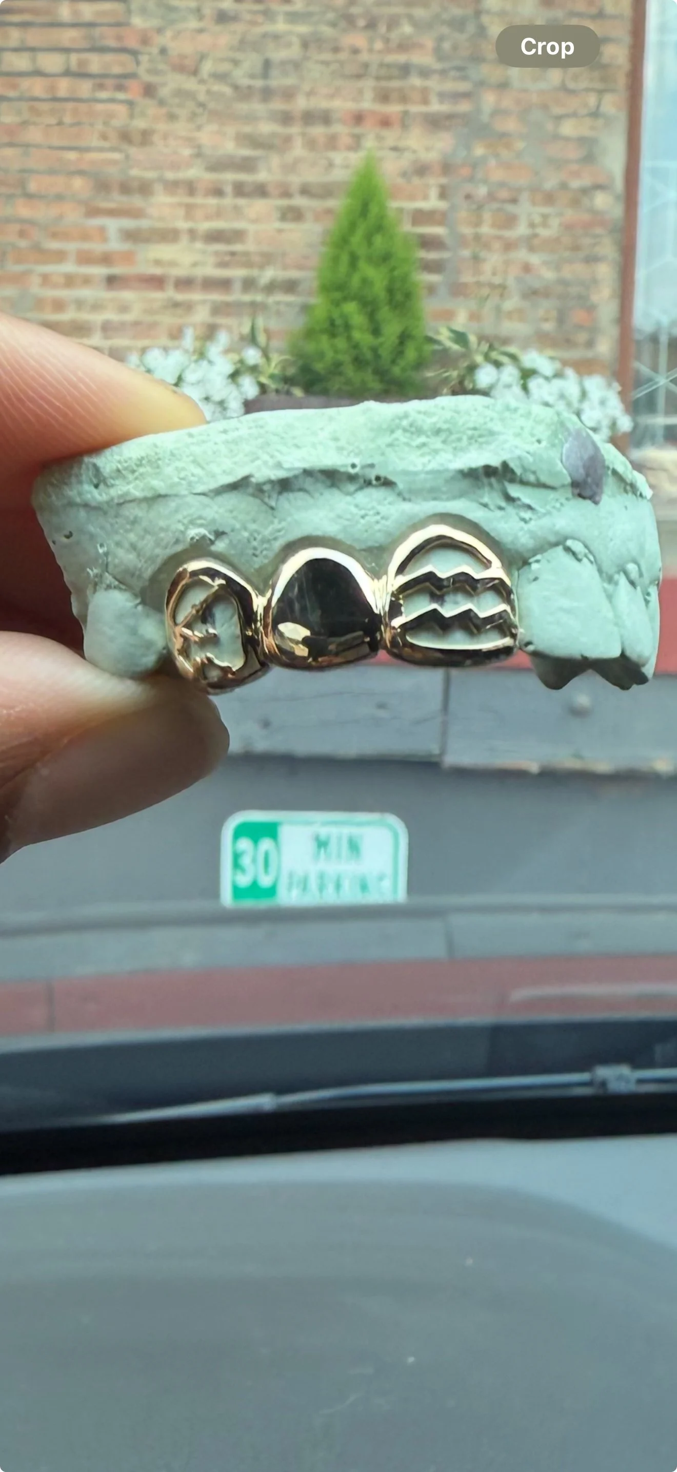 A hand holding a dental impression with gold teeth in front of a car's windshield with a green street sign reading '30 W 14th Place. on the south side of Chicago.
