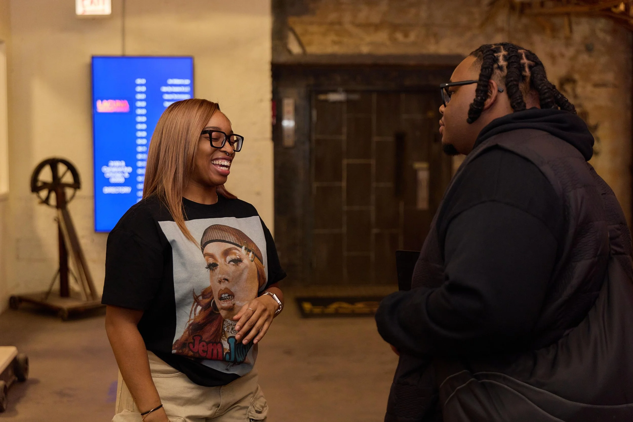 A woman with glasses and blonde hair laughing while talking to a man with glasses and braided hair in a black jacket in an indoor setting at Jem Junkies Tooth Gems and  Custom Grillz in Chicago 