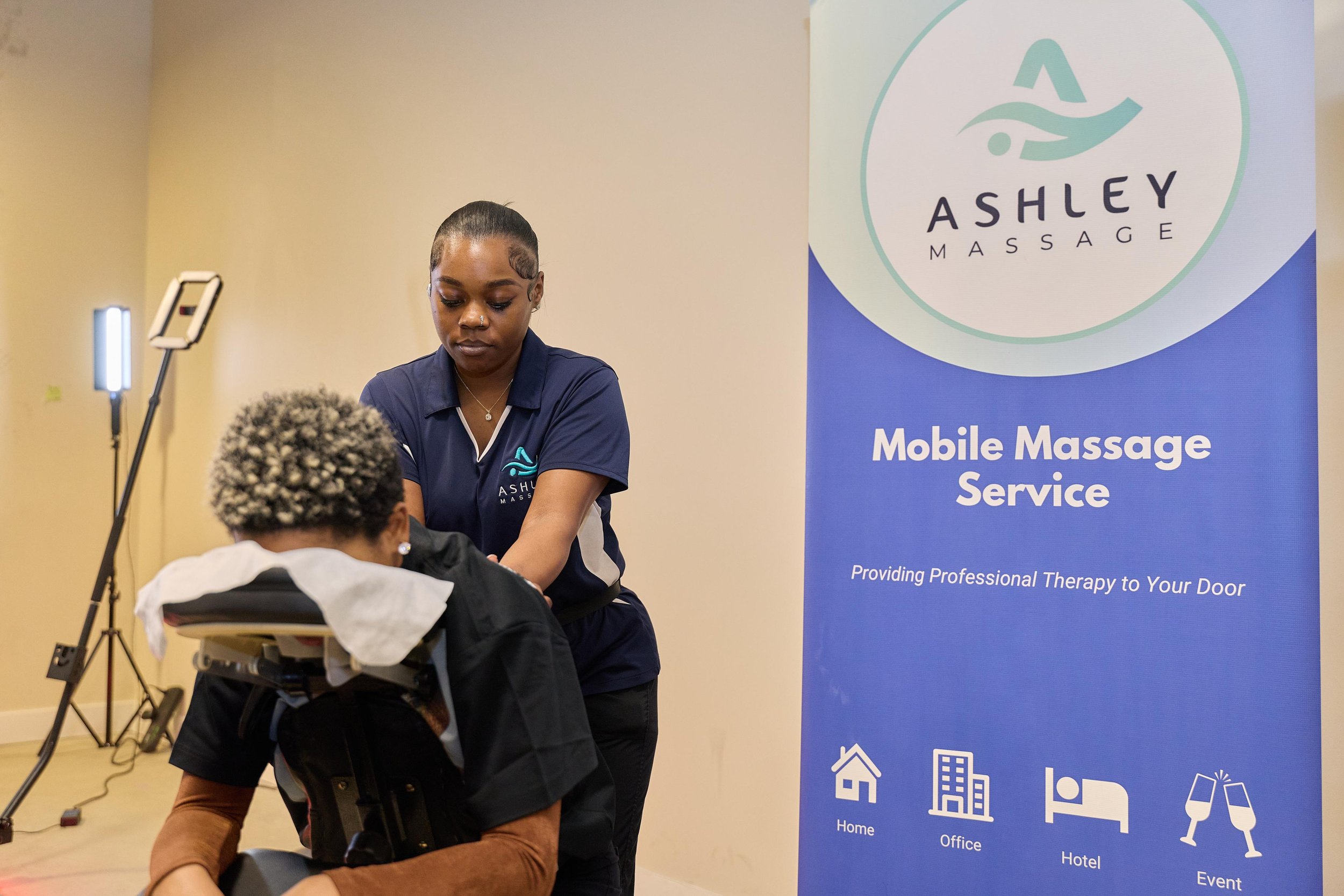 A massage therapist providing a massage to a client in a professional setting. The therapist is wearing a navy blue uniform with a logo that says 'Ashley Massage' and standing beside a seated client with short, curly, gray hair. A large blue promotio
