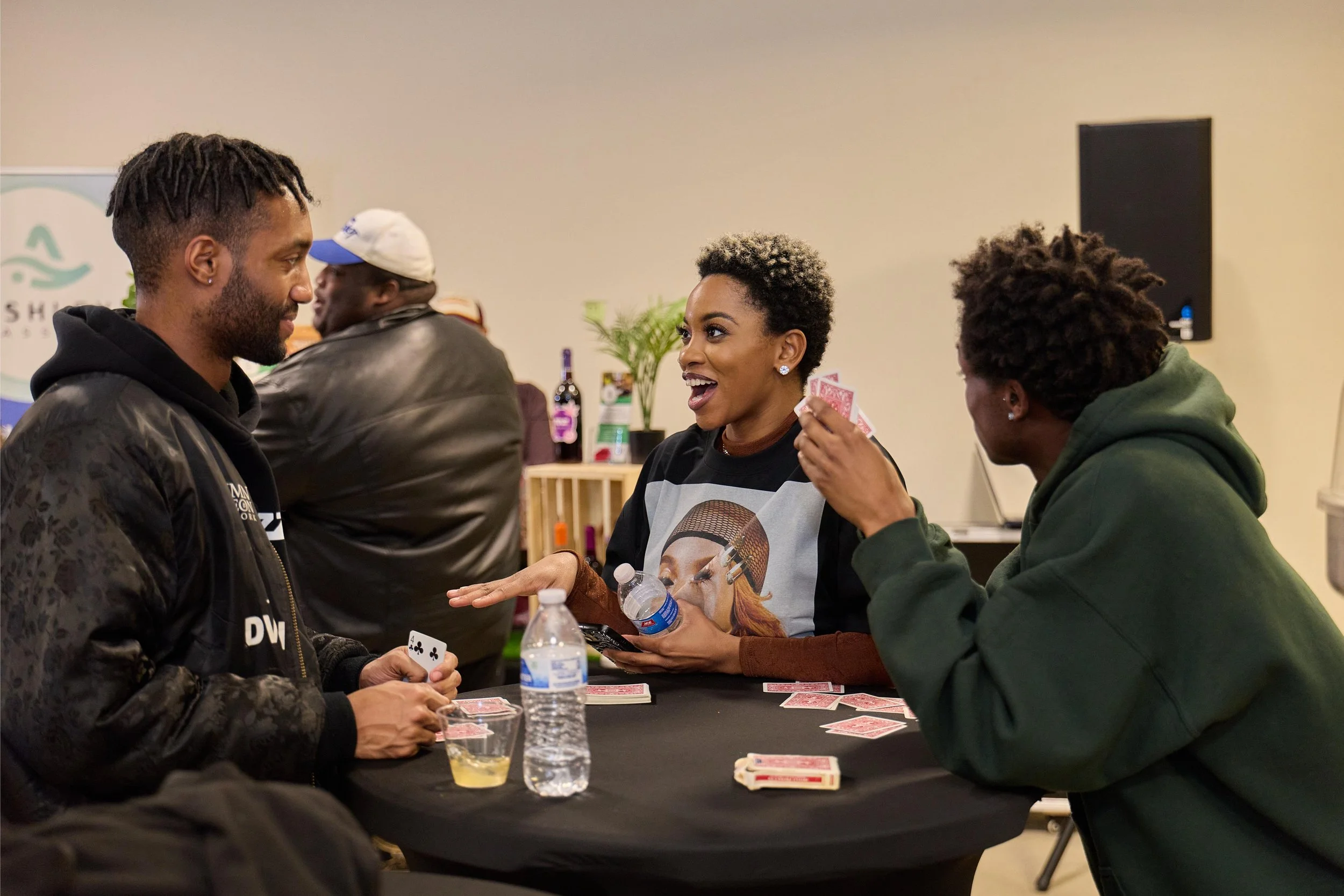 Three people playing cards at a table, smiling and talking, with one person holding cards and the other two showing cards, in a social setting.