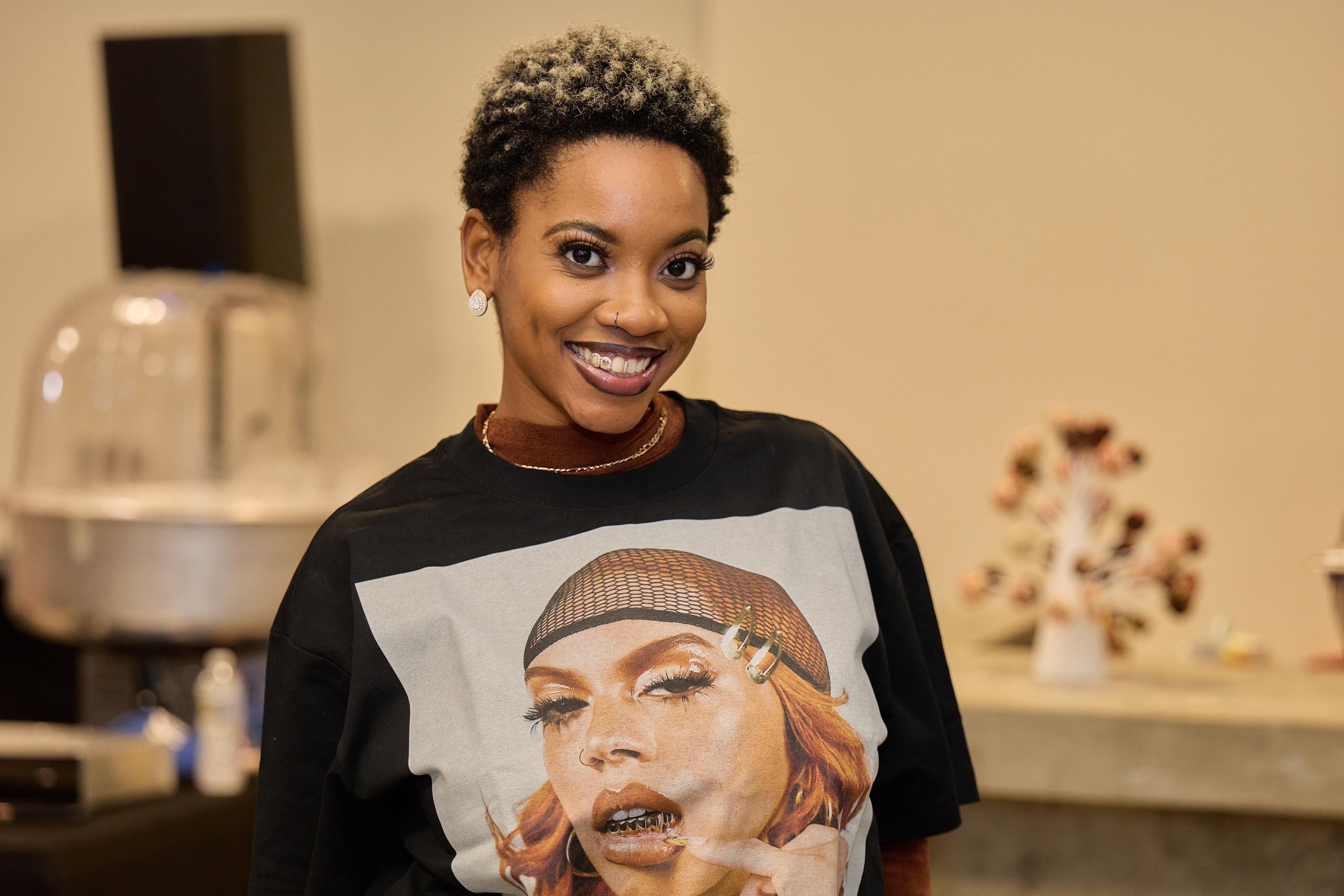 A smiling woman with short curly hair, earrings, and a nose piercing, wearing a black T-shirt with a graphic of a woman at Jem Junkies Tooth Gems and  Custom Grillz in Chicago 