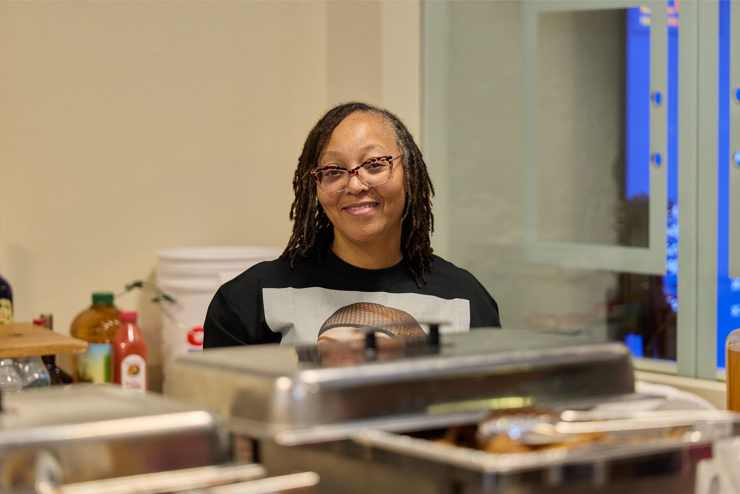 A woman with glasses and dreadlocks smiling in a kitchen or food service area. There are food items and condiments on the counter at Jem Junkies Tooth Gems and  Custom Grillz in Chicago 
