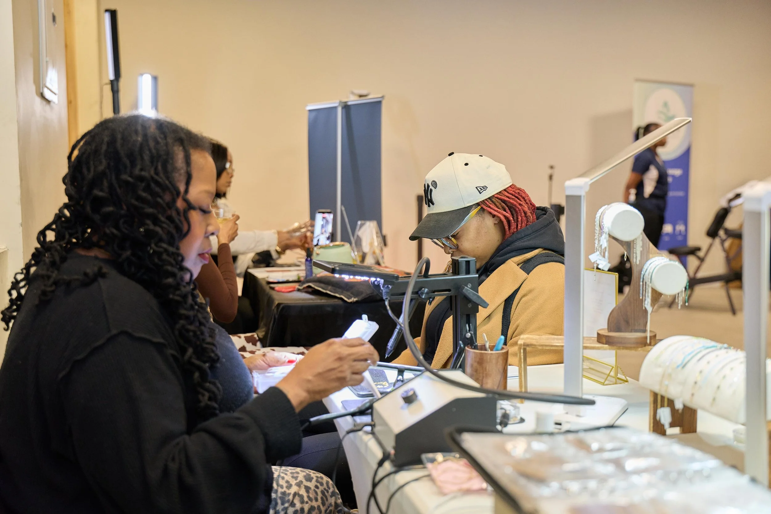 People sitting at a table, one using a phone, with jewelry display stands on the table.