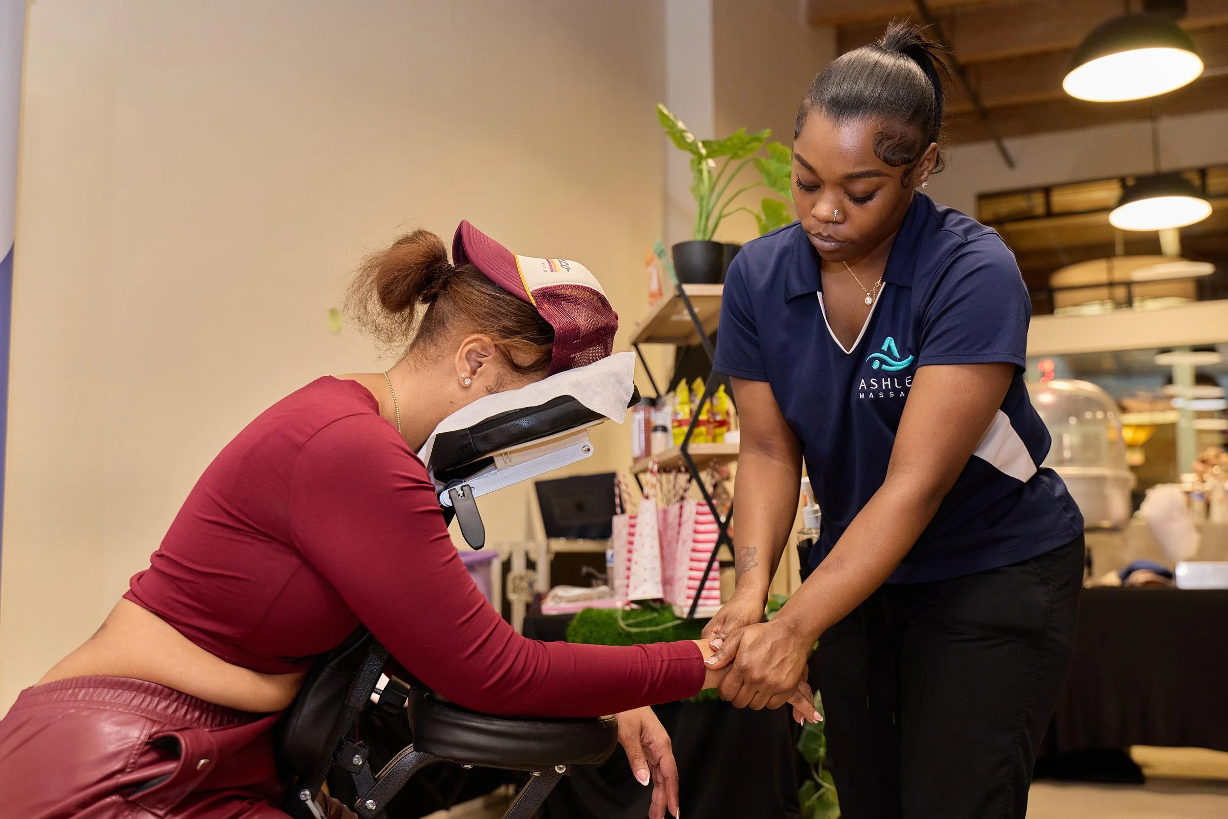 A woman receiving a massage while seated, with her head resting on a massage chair, as another woman stands beside her applying the massage, in a spa or massage therapy setting at Jem Junkies Tooth Gems and  Custom Grillz in Chicago 
