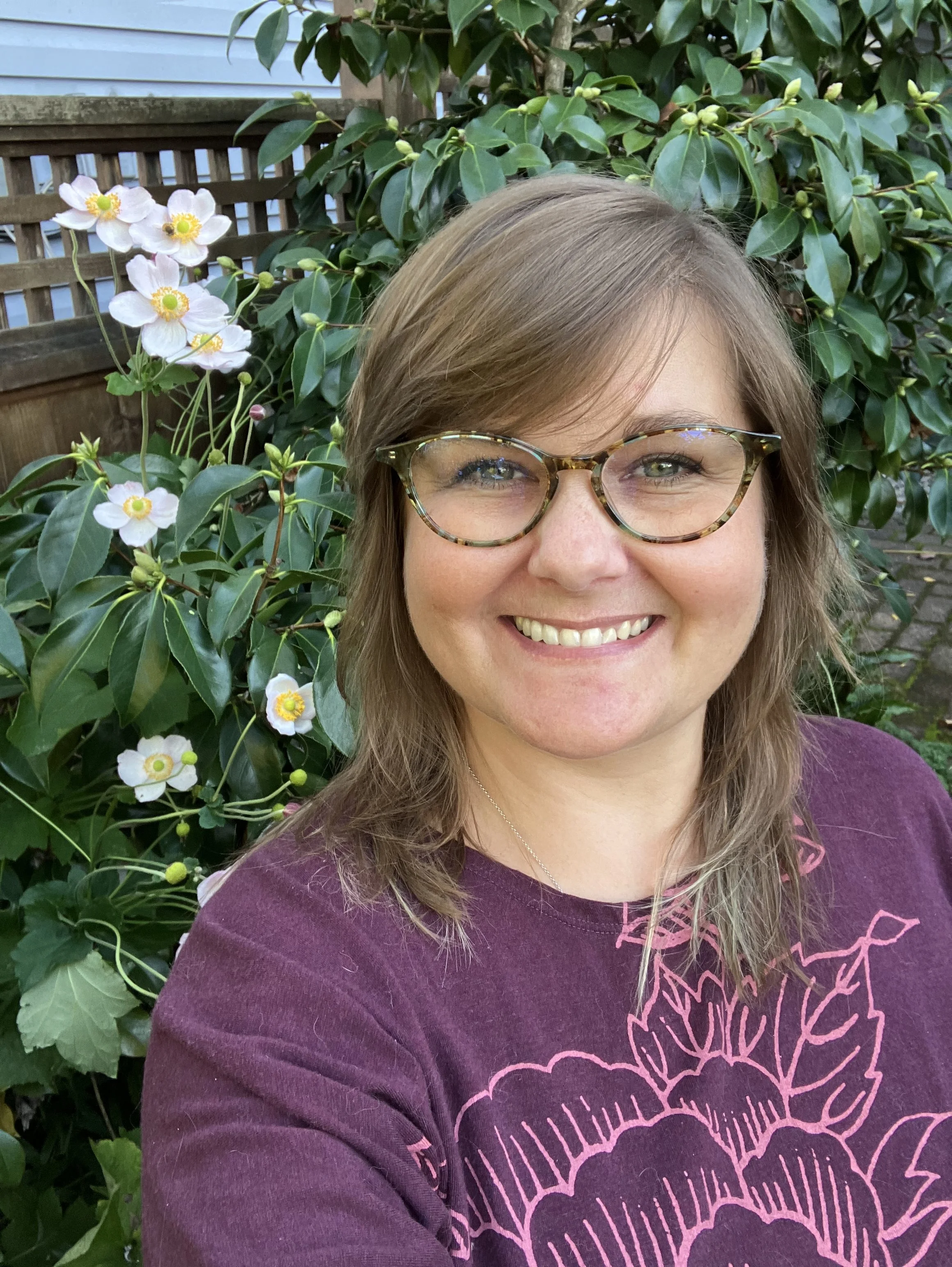 Smiling person with glasses and brown hair taking a selfie in front of a garden with white flowers and green leaves.