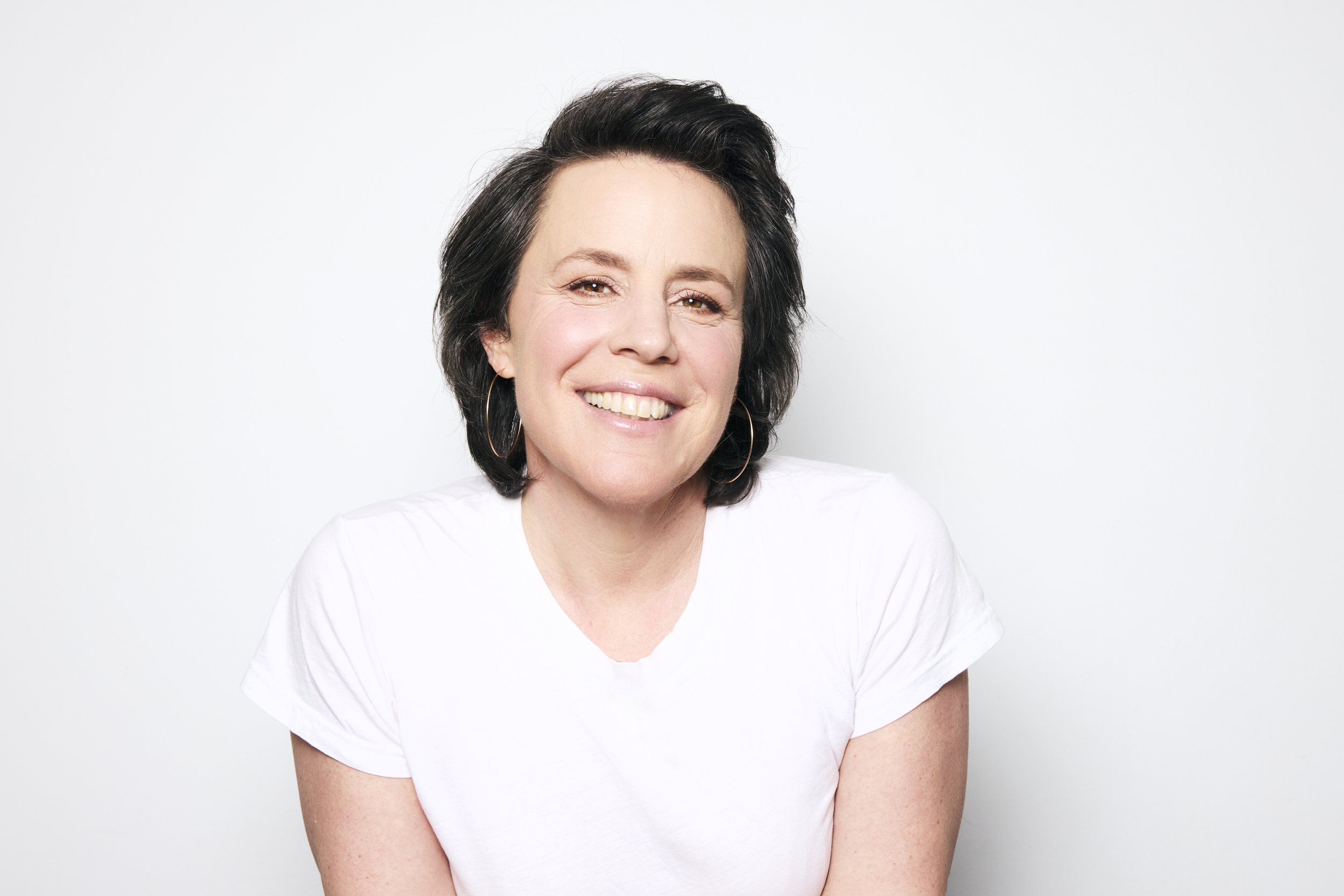 A woman with short dark hair smiling and wearing a white t-shirt against a plain white background.