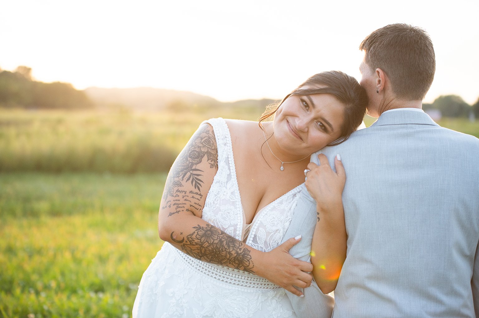 Bride leaning on groom's shoulder in a field at sunset, smiling, wearing a white lace dress with tattoos on her arm.