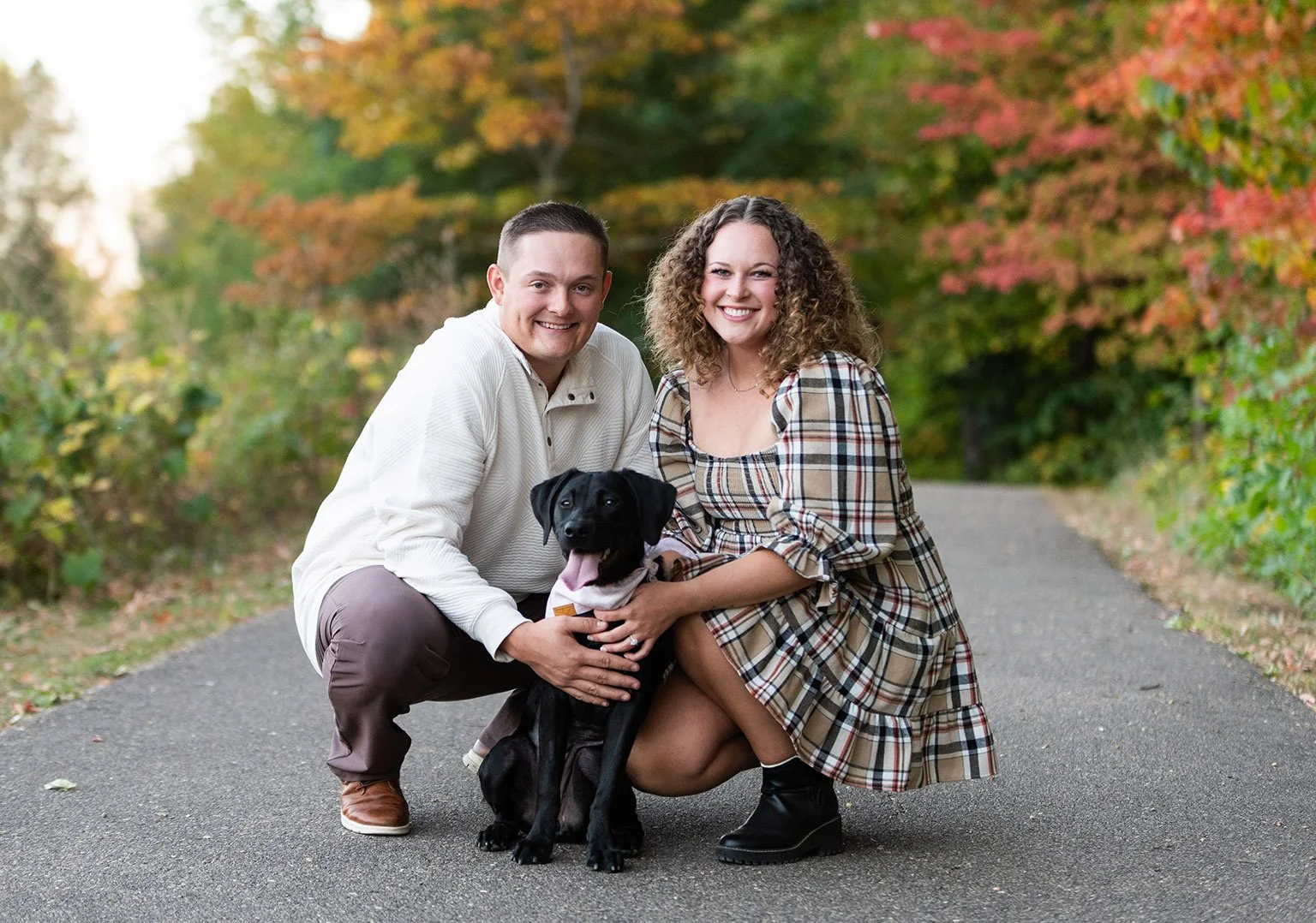 A man and woman crouching on a path with a black dog, surrounded by autumn foliage.
