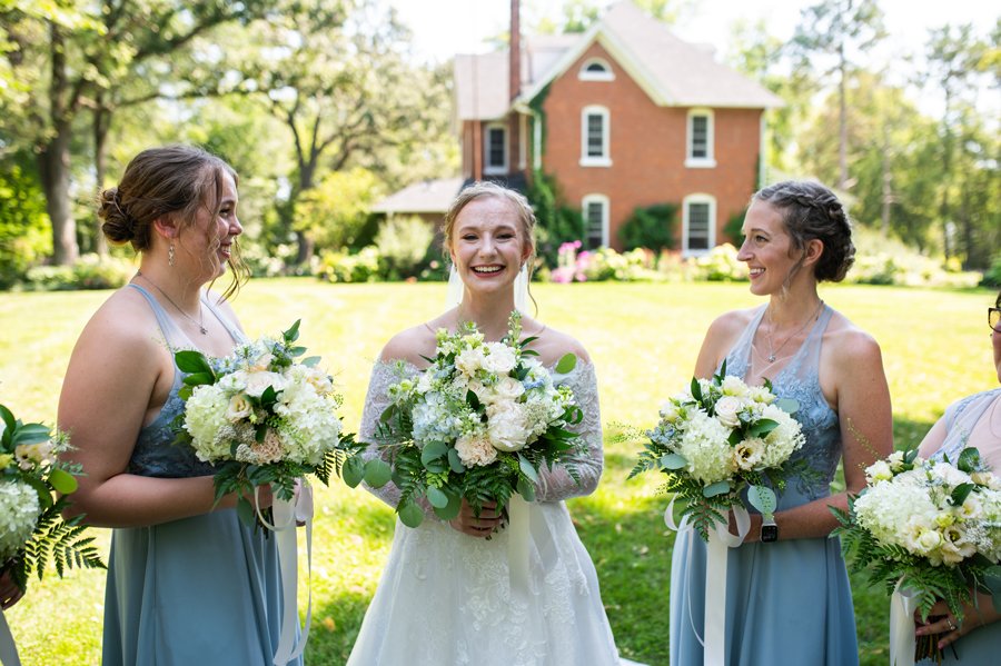 Bride and bridesmaids holding bouquets, standing outdoors with a brick house in the background.