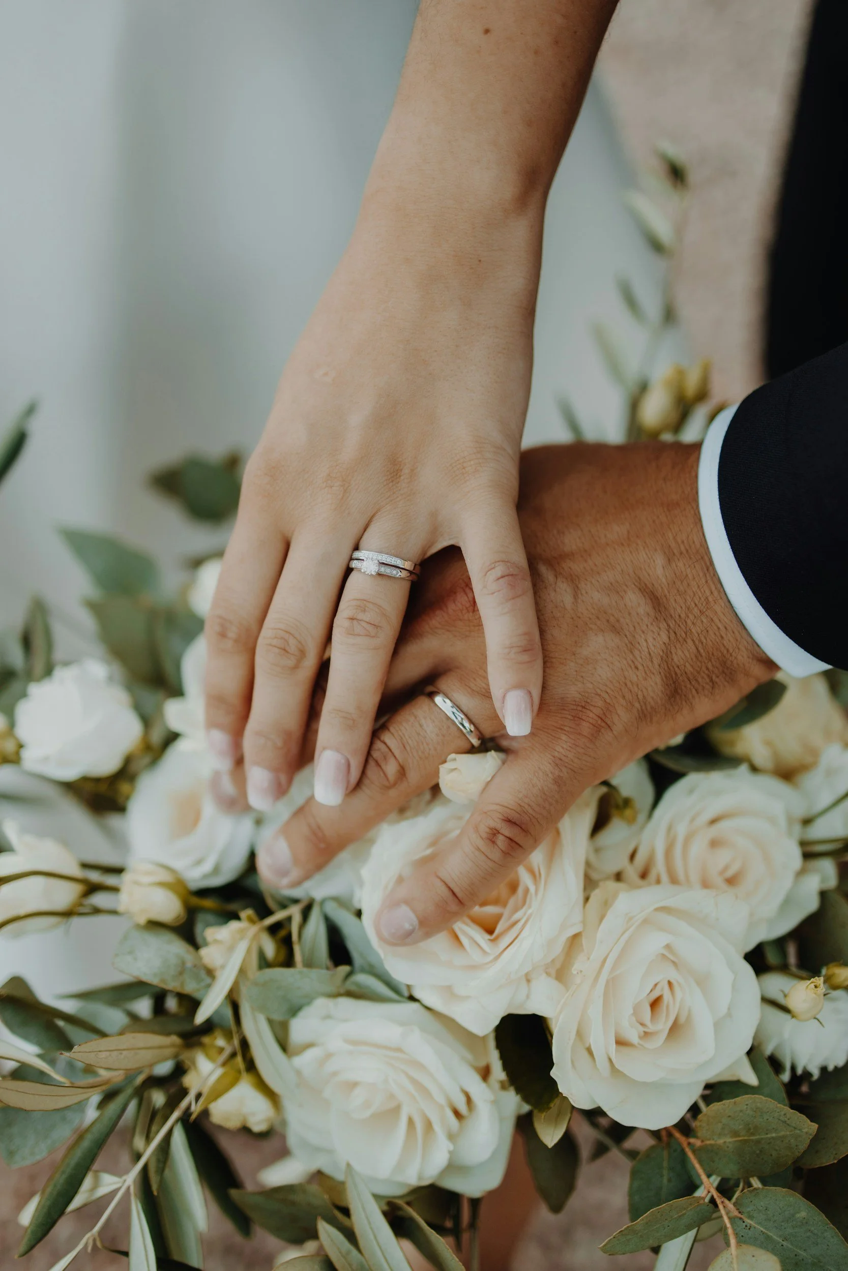 Close-up of a couple holding hands over a bouquet of white and cream roses with green leaves, showing wedding rings on their fingers.