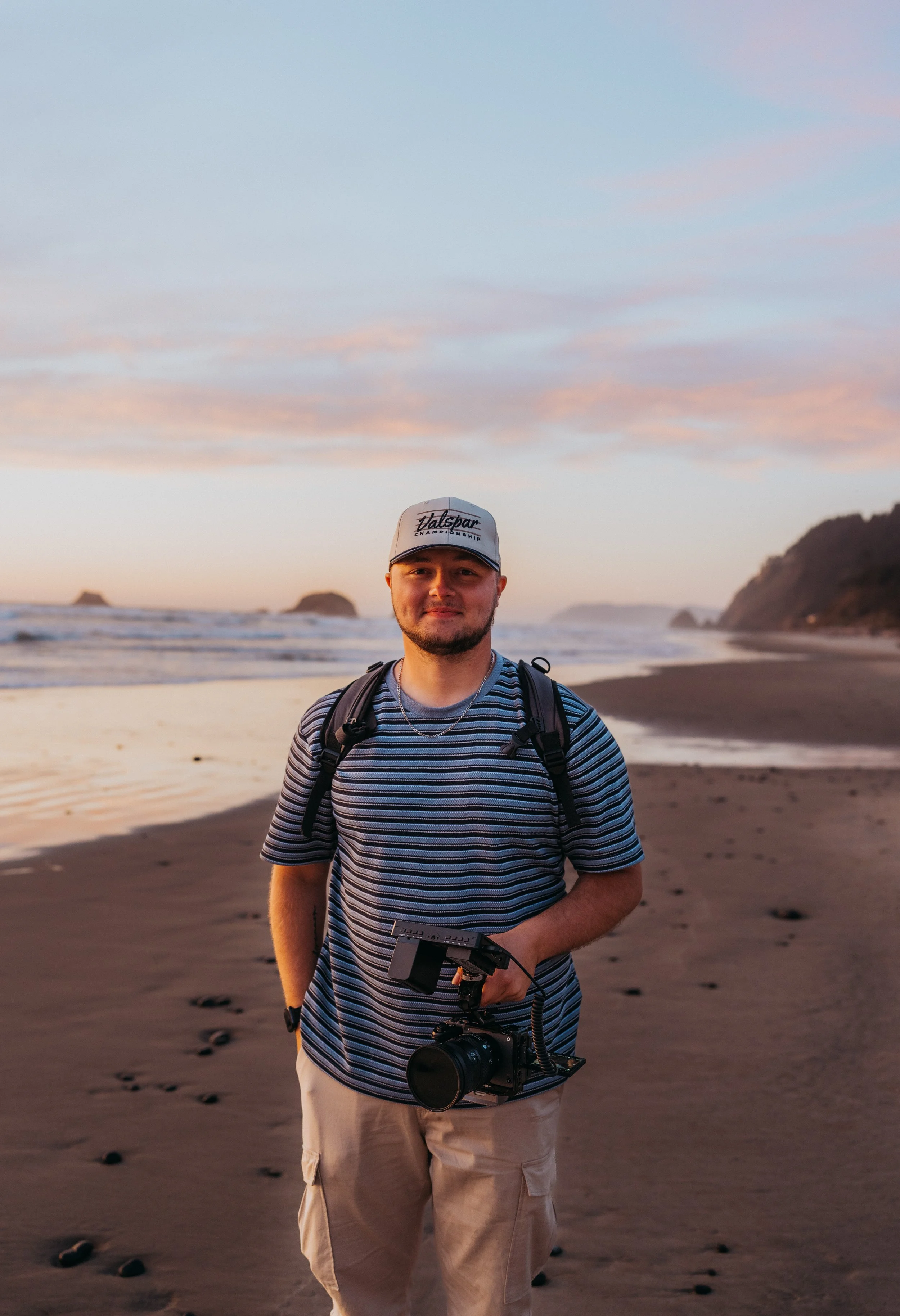 A man with a camera on a beach during sunset, wearing a striped shirt, beige cargo shorts, a cap, and a backpack.
