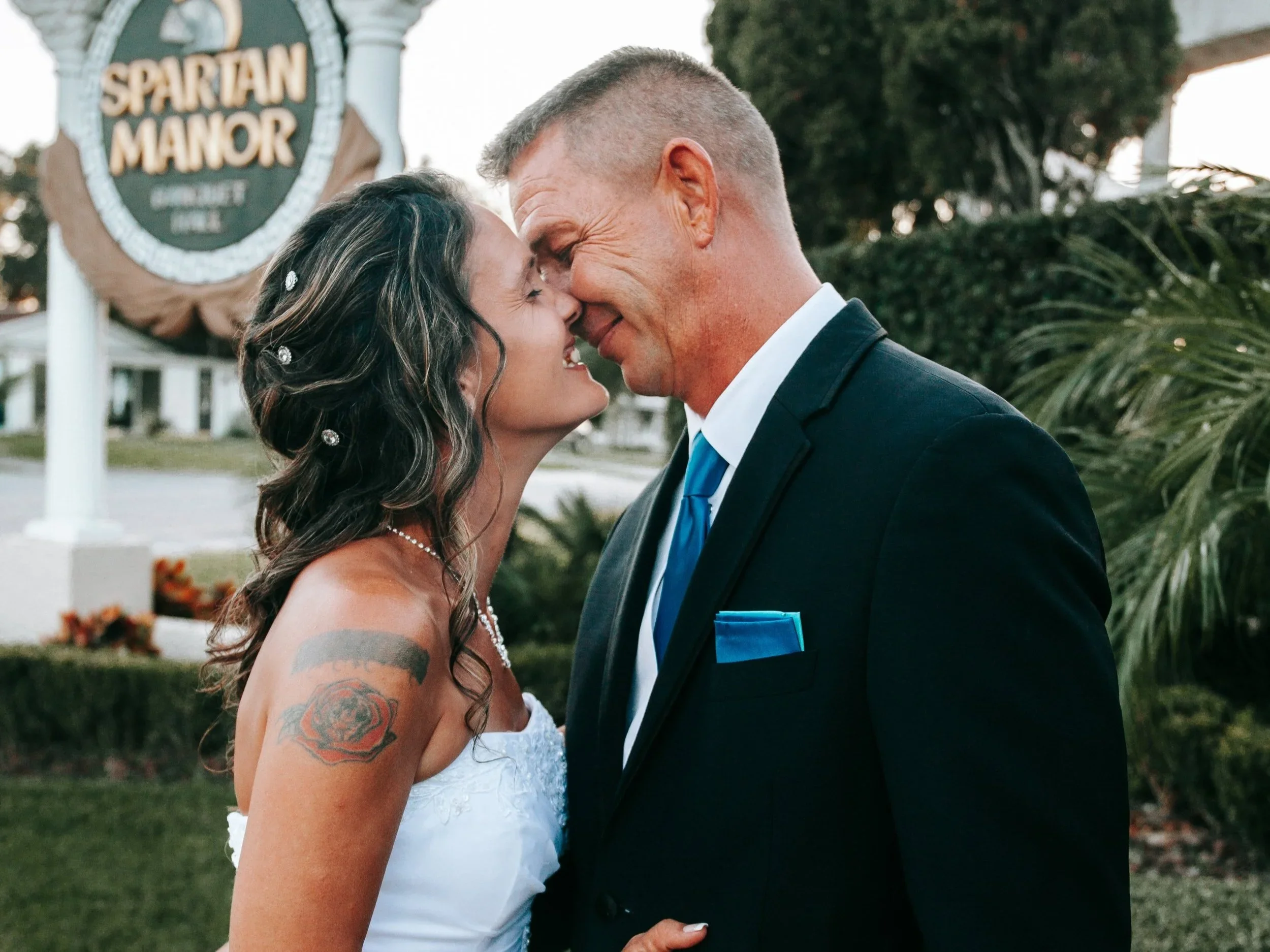 A bride and groom share a close, intimate moment with their foreheads touching, outdoors in front of a sign that reads 'Spartan Manor.' The bride has dark, curly hair decorated with pearls, visible tattoos on her arm, and is wearing a white lace wedd