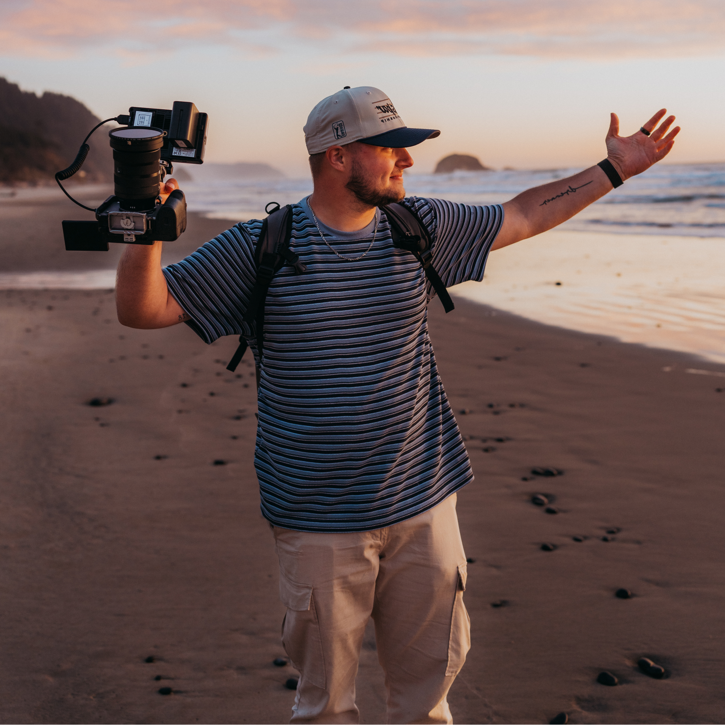 A man is standing on a beach during sunset, holding a camera with a large lens in his right hand and waving with his left hand. He is wearing a striped t-shirt, khaki shorts, a white and navy cap, and has a backpack. There are rocks and water in the background, and footprints are visible on the sand.