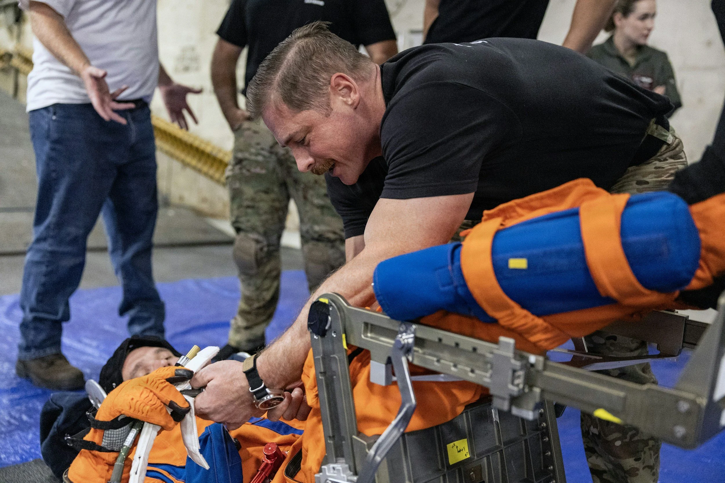 A man lying on a medical stretcher, connected to medical equipment, appears to be receiving emergency care. Others are standing around in what looks like an emergency or training setting.