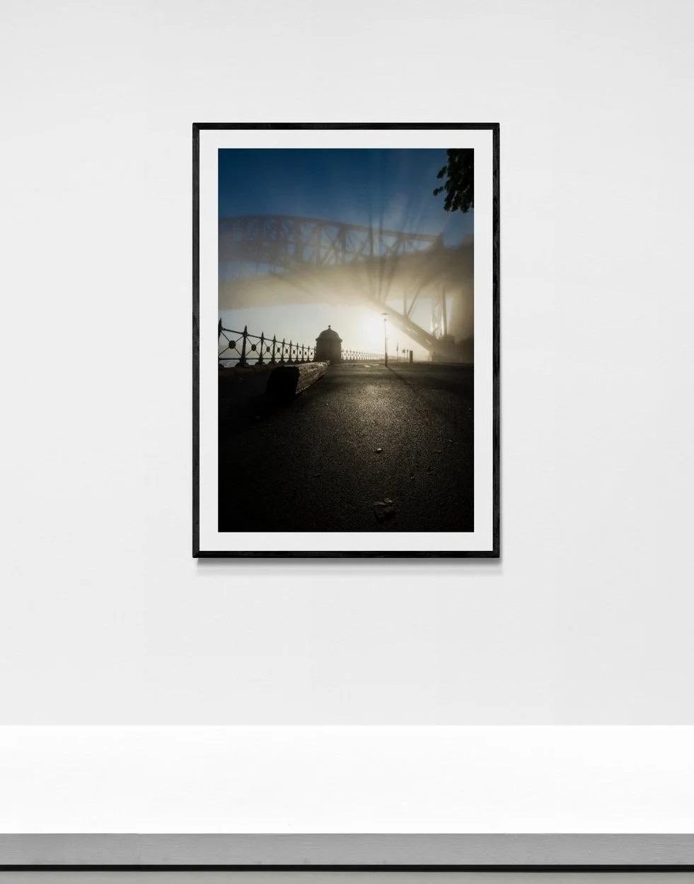 A framed photograph of a bridge taken during sunrise or sunset with sun rays in the background, positioned on a white gallery wall.