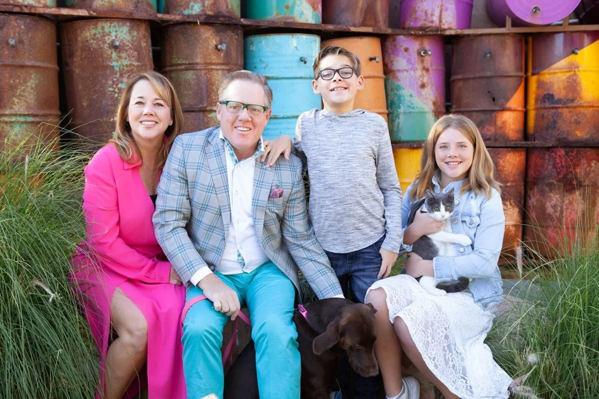 A family of four with two children and two pets sitting outdoors in front of a stack of colorful, rusty barrels. The mother is wearing a bright pink dress, the father a plaid blazer and glasses, the son in a gray hoodie, and the daughter in a white lace dress holding a black and white cat. A brown dog is seated on the grass in front of the family.
