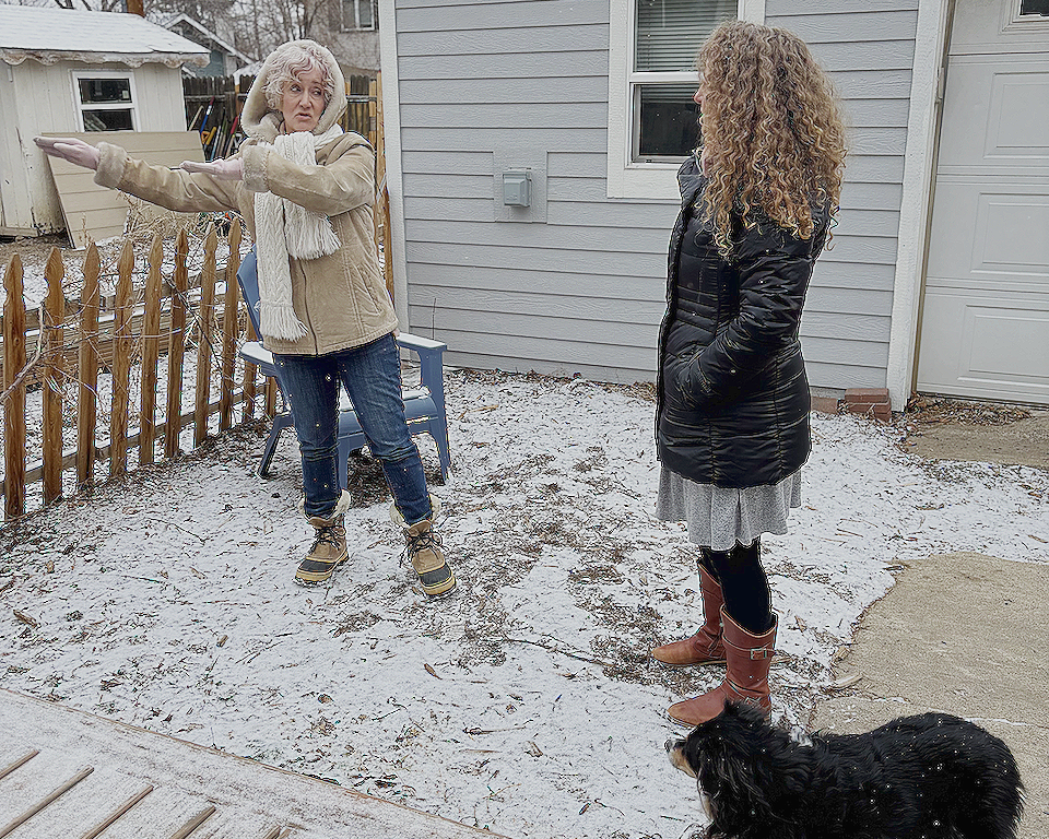 Two women and a dog outside in a snowy backyard. One woman, with gray hair, is gesturing to the side, while the other woman has curly hair, is standing with hands in her coat pockets, and is watching her. The dog, a black and brown spaniel, is on the ground near them.