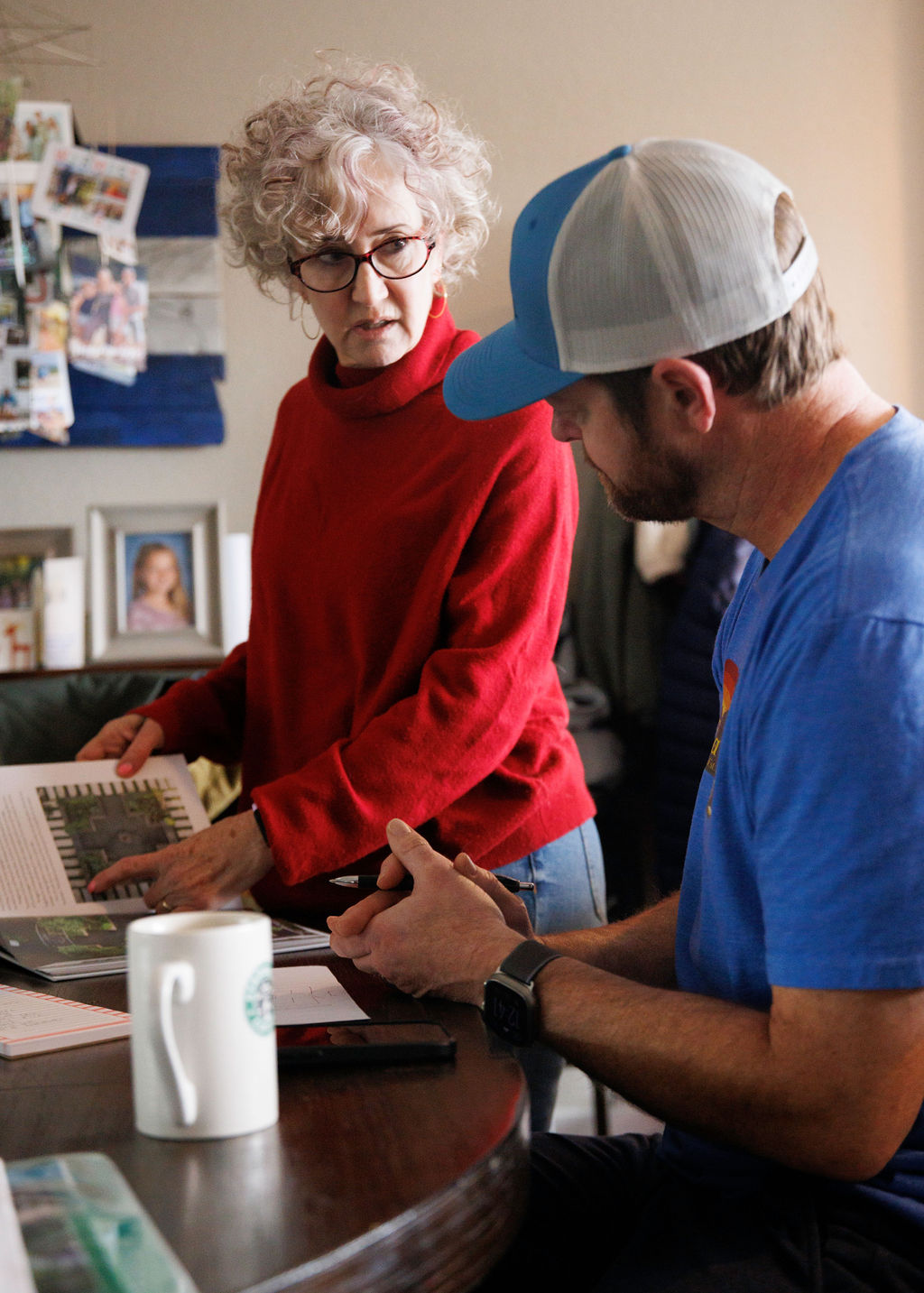A woman with curly gray hair and glasses, wearing a red sweater, is talking to a man with a beard, wearing a blue t-shirt and a white and blue cap. They are in a room with a table that has a coffee mug, a smartphone, and documents, and a picture of a young girl on a shelf behind them.
