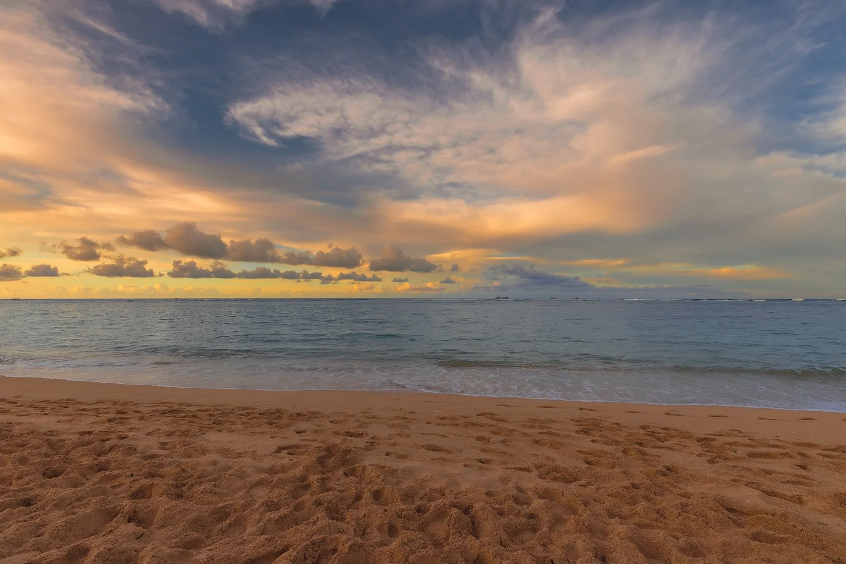 A sandy beach with gentle waves and an expansive sky with scattered clouds during sunset.
