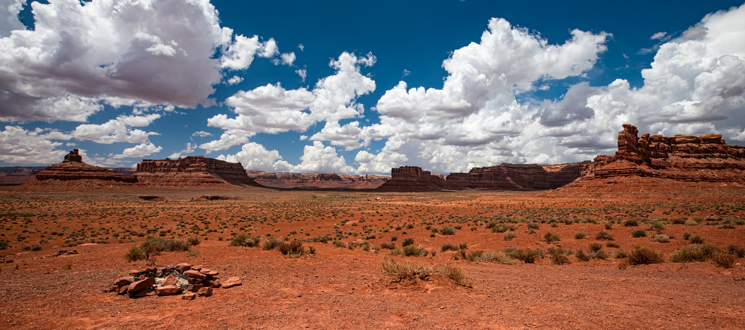 A vast desert landscape with orange reddish soil, sparse green bushes, large rock formations and mesas in the distance under a partly cloudy blue sky.