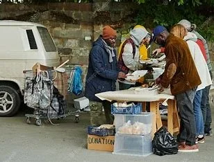 Group of people at a street donation table with a donation box, near parked cars and a shopping cart.