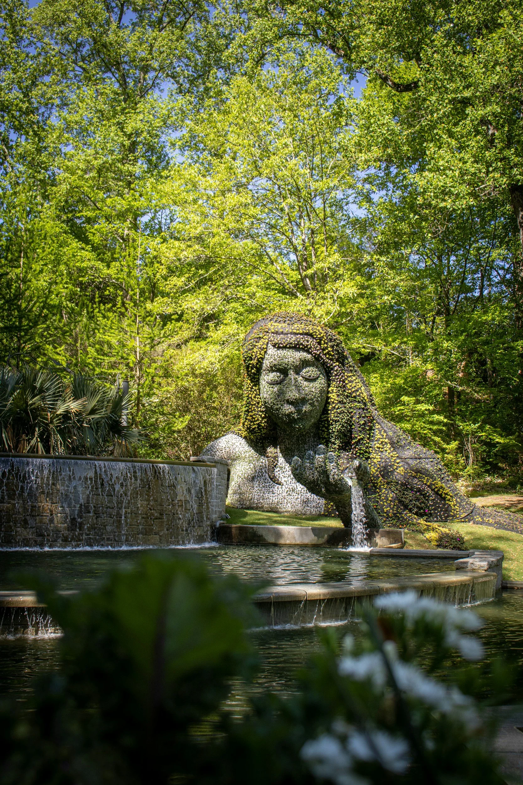 A large outdoor sculpture of a woman's face and upper body, made of stones or mosaic, with water flowing from her hand into a pool, surrounded by lush green trees and plants.