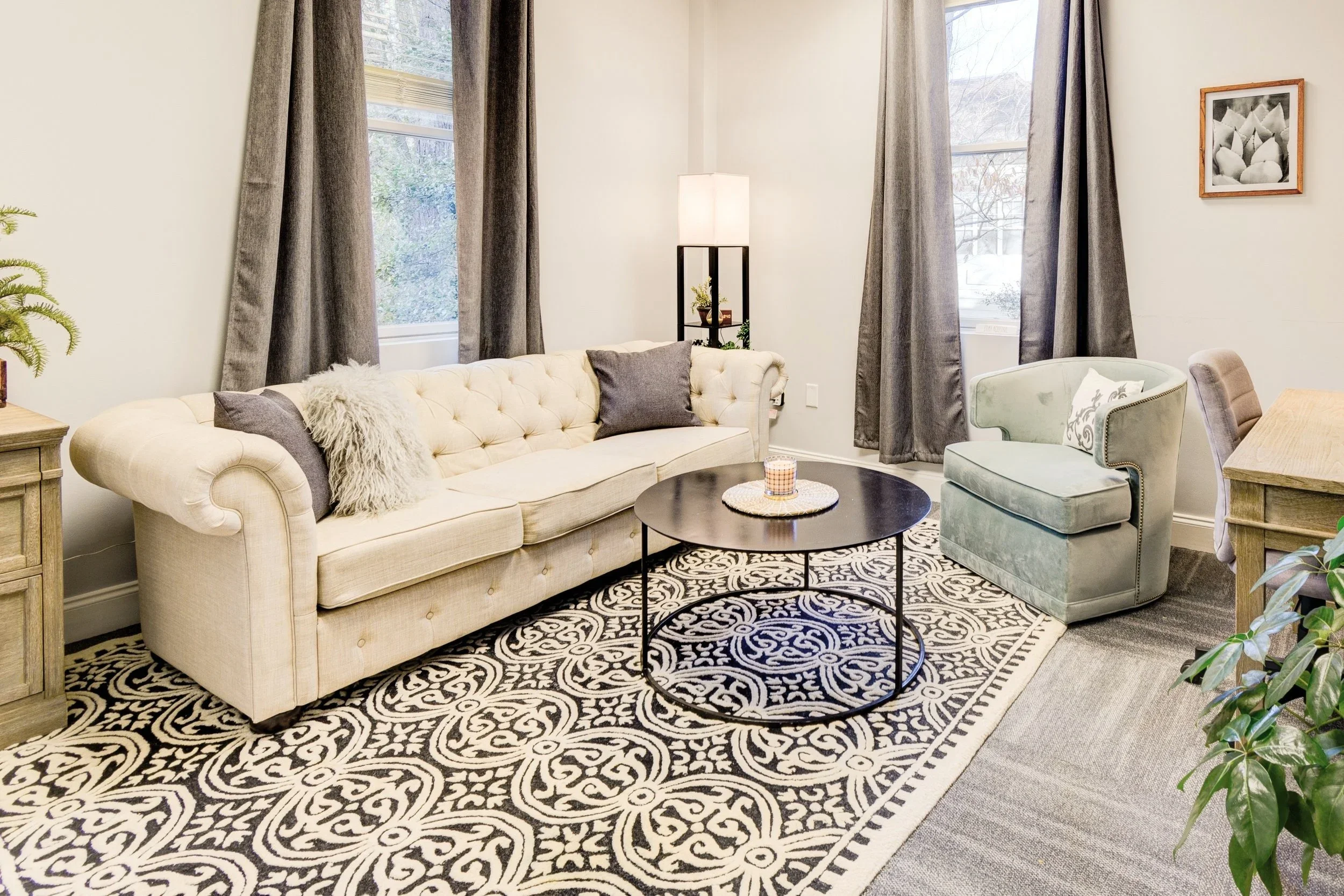 Living room with cream-colored tufted sofa, patterned black and white rug, round black coffee table, light green armchair, side tables, gray curtains, and framed black and white artwork on wall.