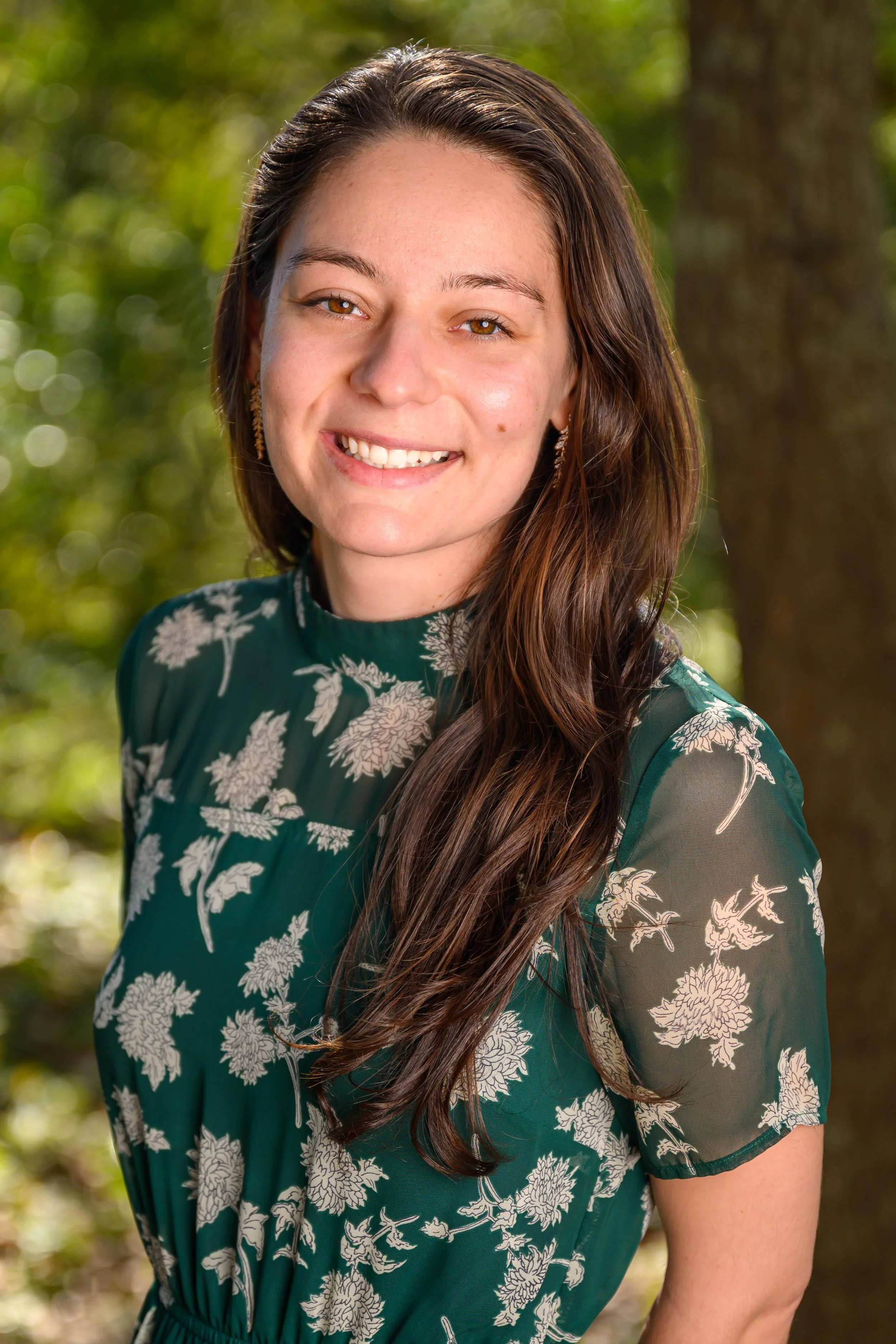 Young woman outdoors with long brown hair, wearing a green floral dress, smiling at the camera with a blurred natural background.