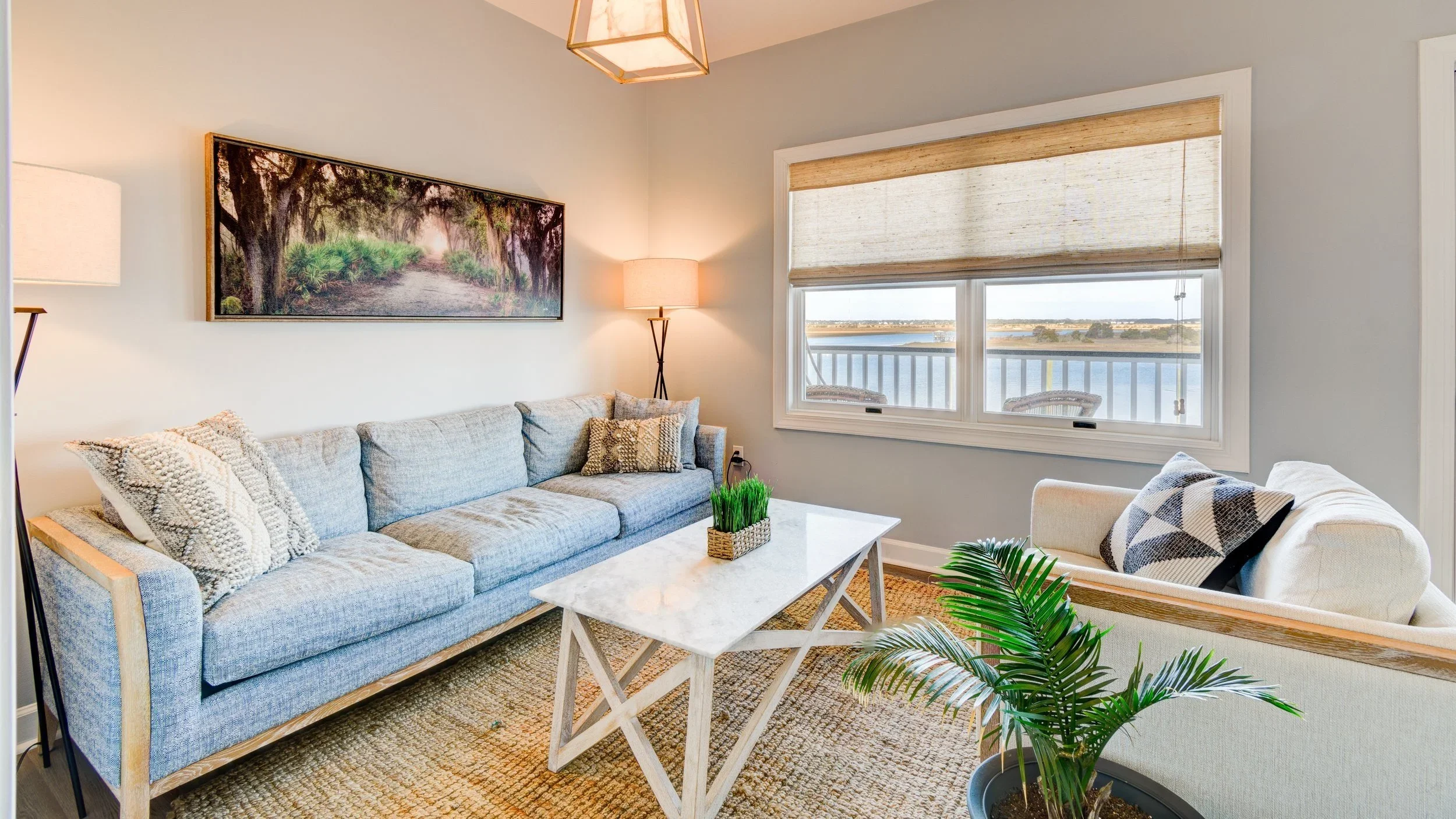 Living room with a large light gray couch, a white marble coffee table, a beige armchair, a window view of water, and decorative plants.