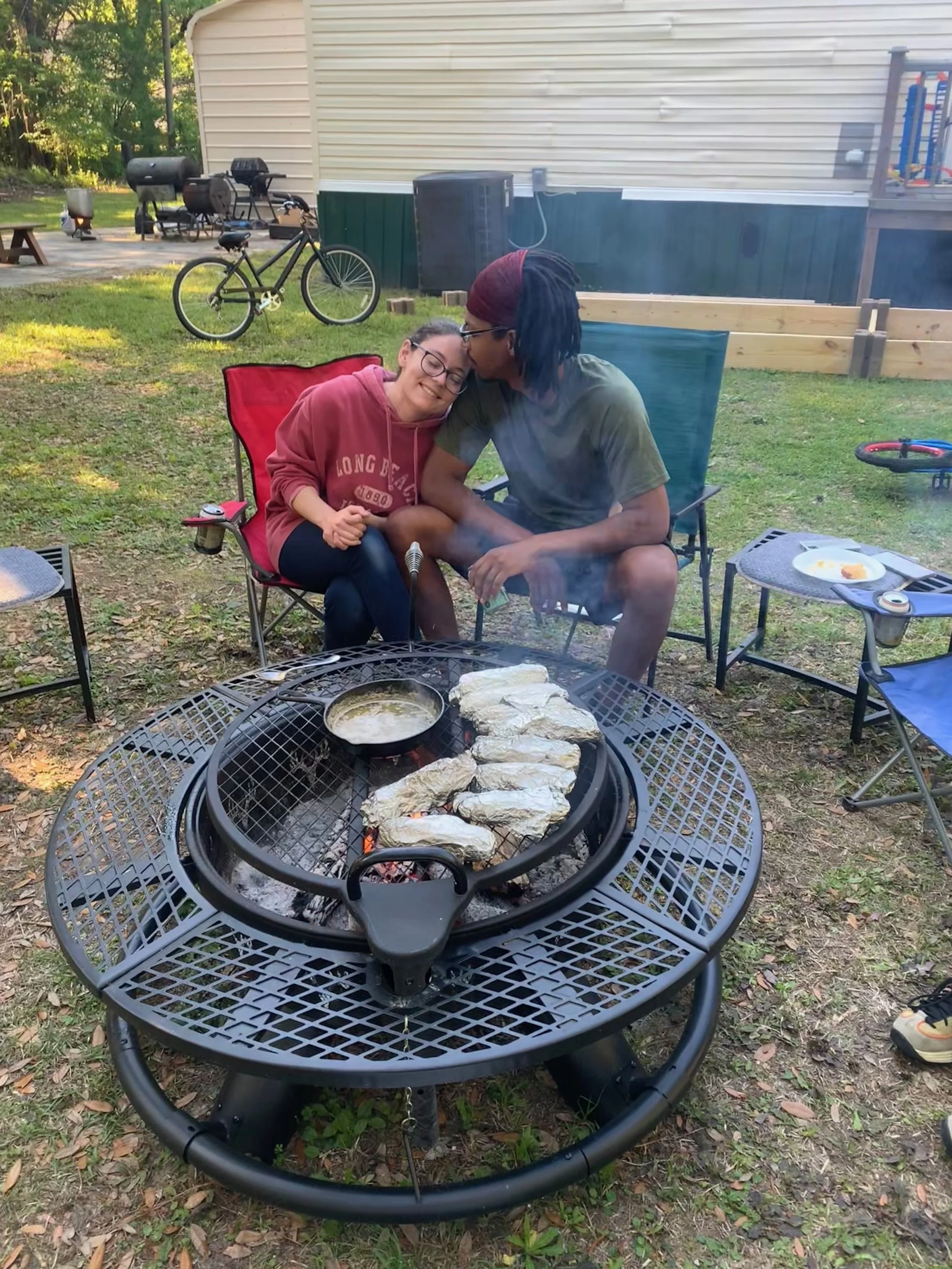 Two people sitting on outdoor chairs, sharing a tender moment, near a barbecue grill with food cooking, surrounded by outdoor furniture and a backyard setting with a house, bicycle, and children’s play area.