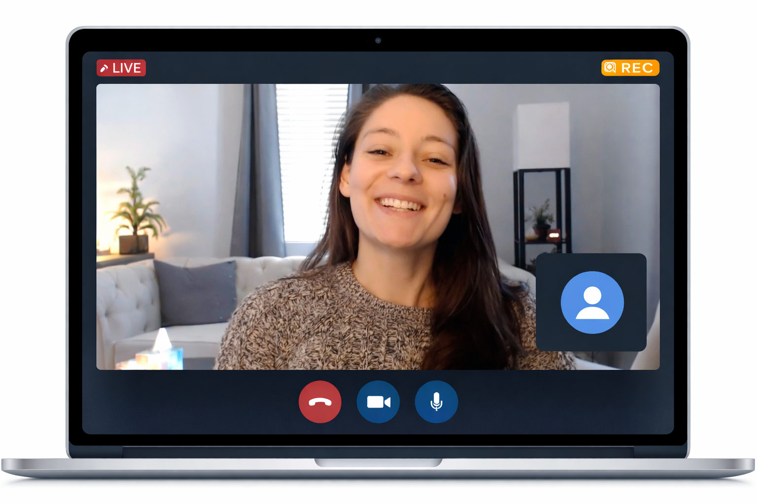 A woman with long dark hair smiling on a video call in a living room with gray walls, a beige sofa with pillows, a window with gray curtains, and a black shelving unit with plants and decorations.