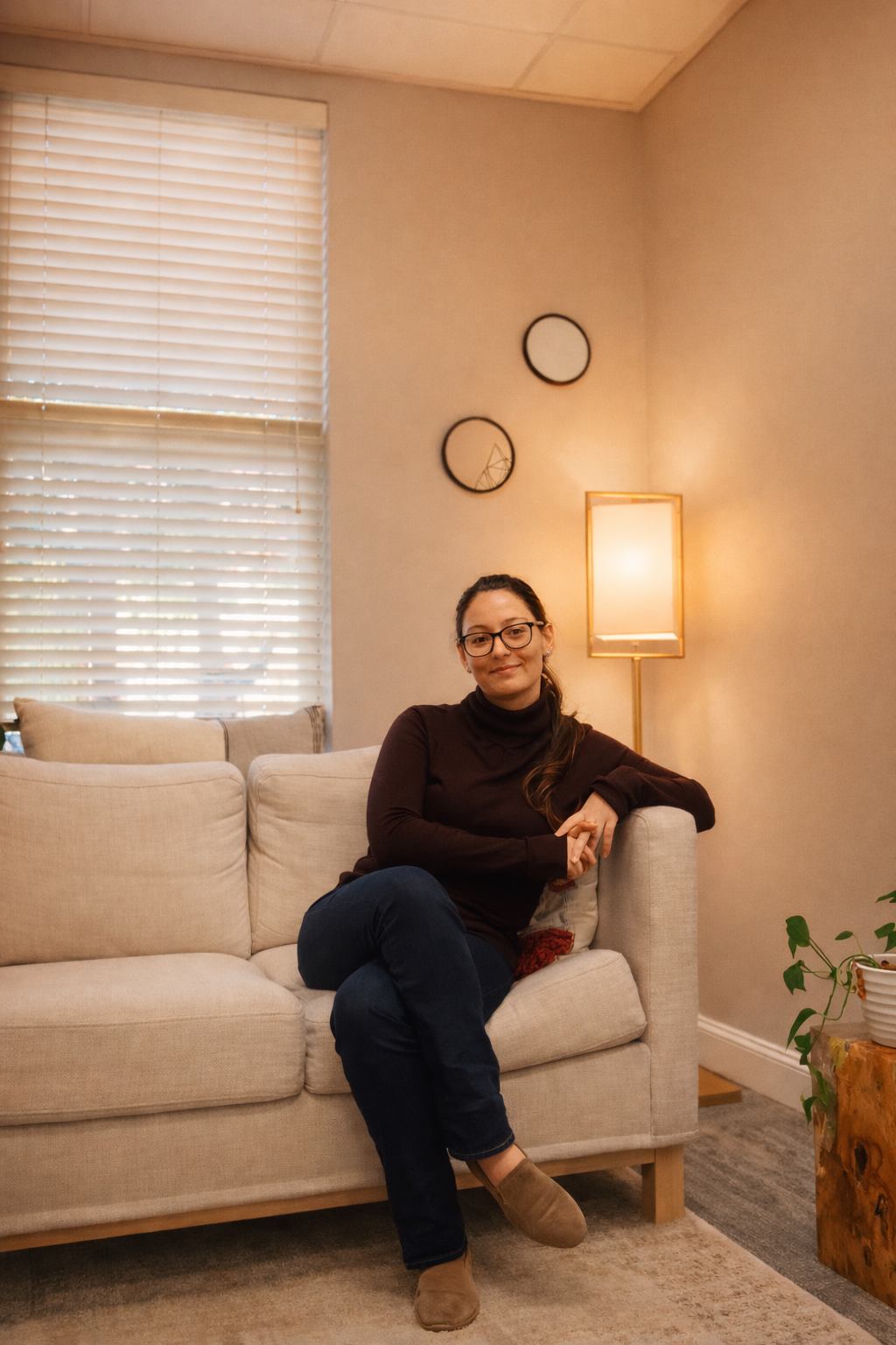 A woman sitting on a beige couch in a cozy living room, with beige walls, a tall floor lamp, decorative wall mirrors, and a potted plant on a wooden side table.