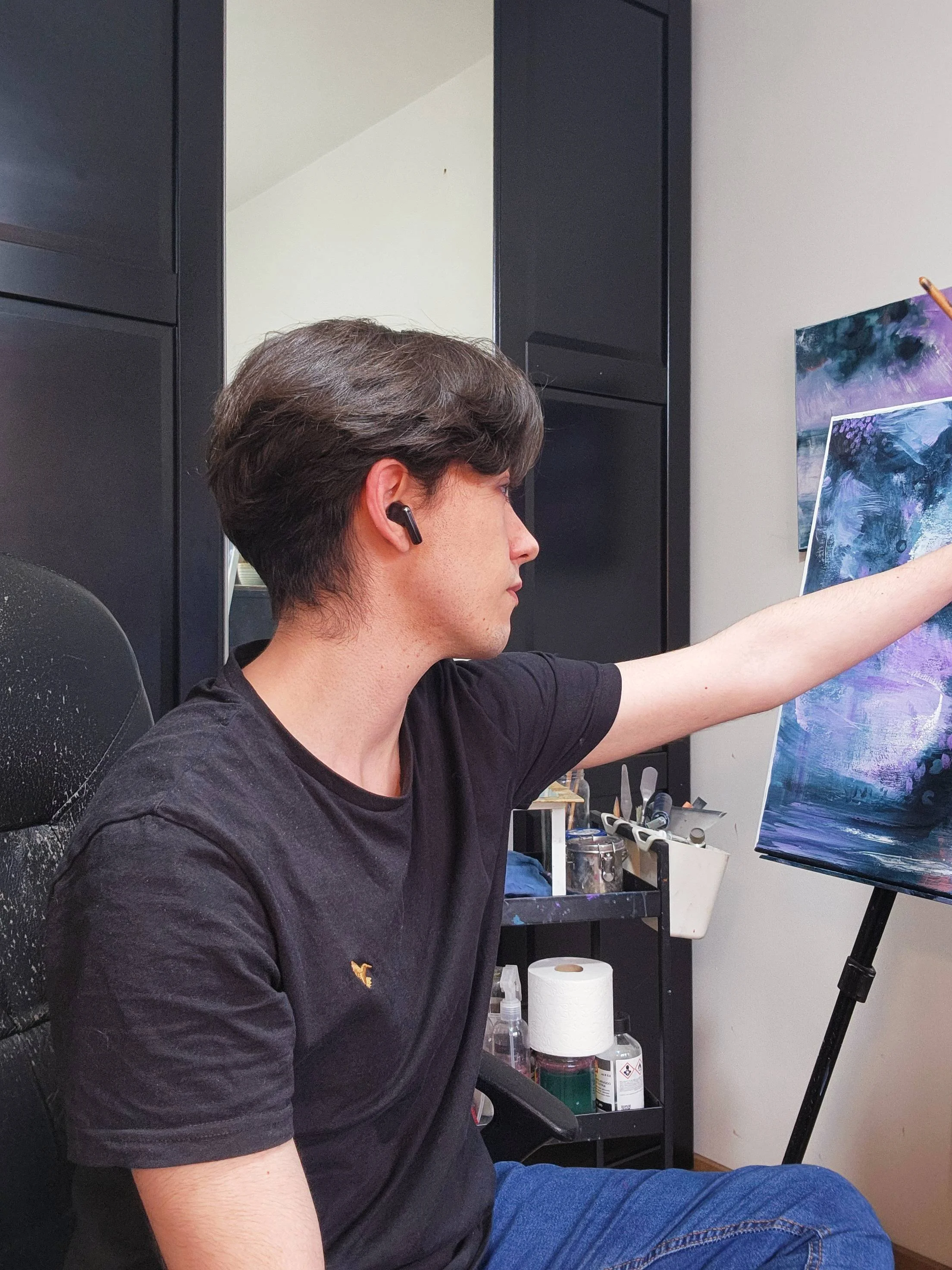 Joven pintando en un caballete, usando audífonos inalámbricos en un estudio con materiales de arte y almacenamiento.