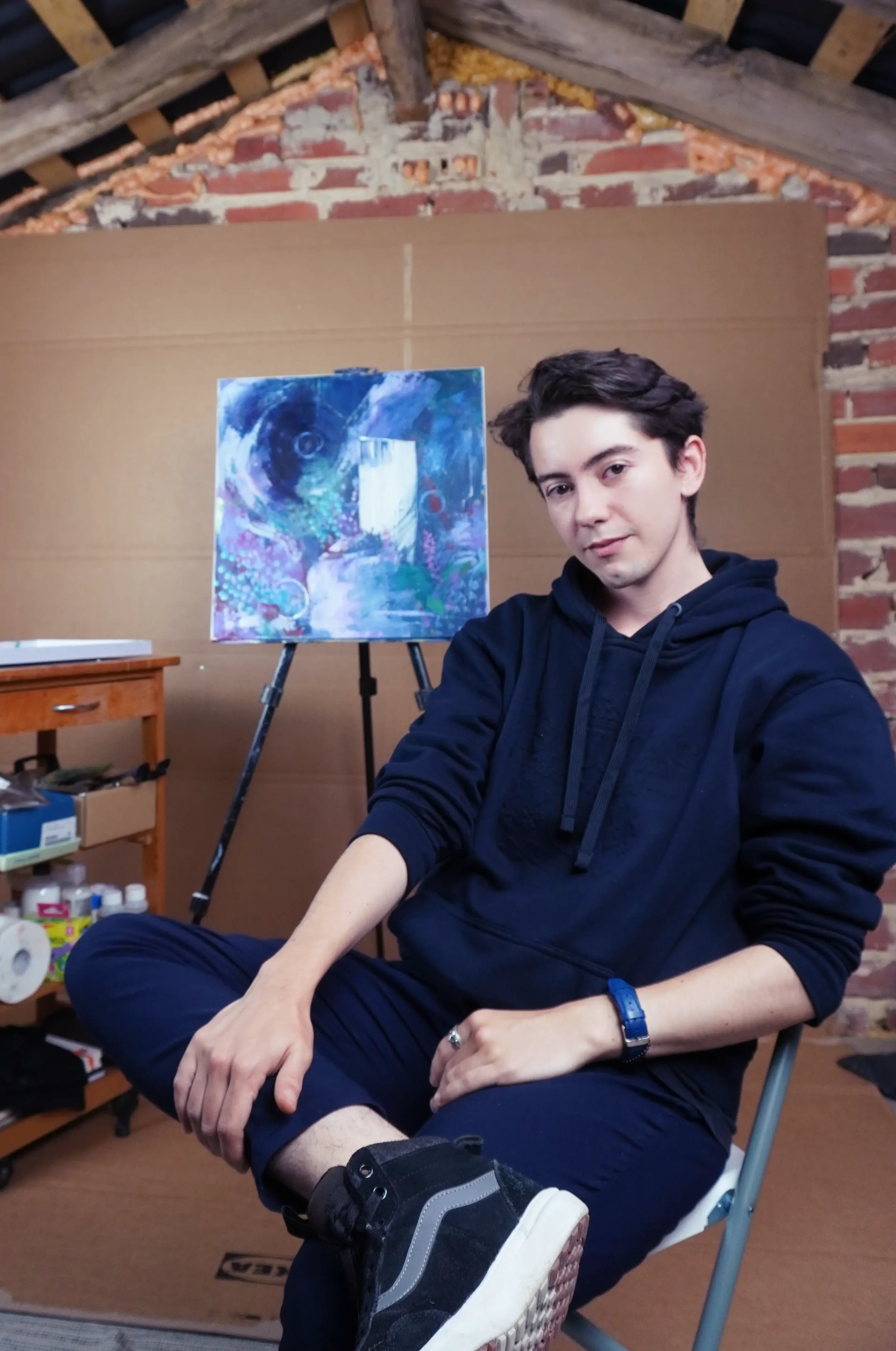 Joven sentado en una silla de plástico con una pierna levantada, con zapatillas deportivas negras, en un taller o estudio de arte que tiene una pintura abstracta en caballete y materiales de arte en una mesa al fondo.