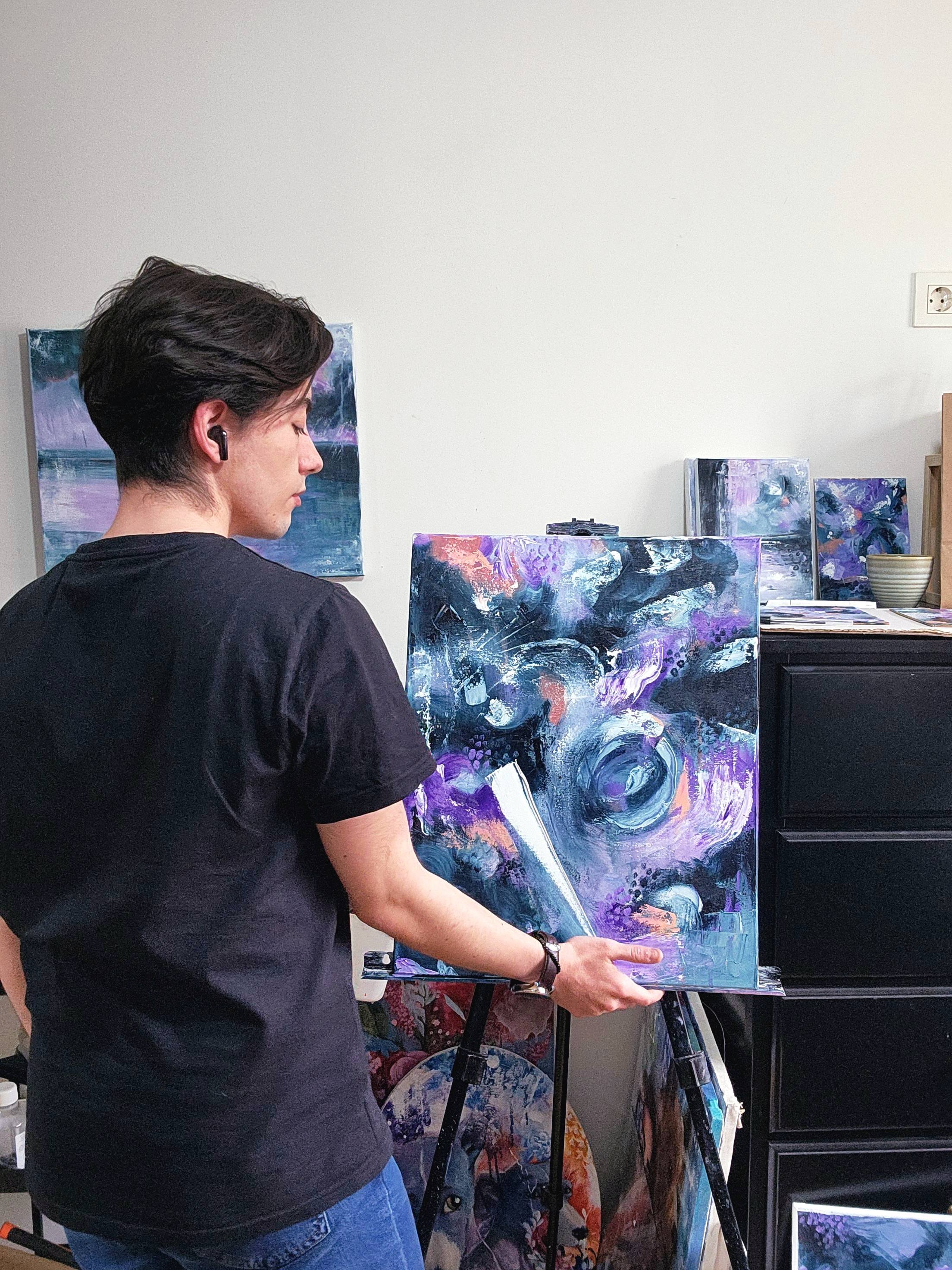 Una persona pintando un cuadro abstracto en un caballete, con otras obras de arte y materiales en el fondo.