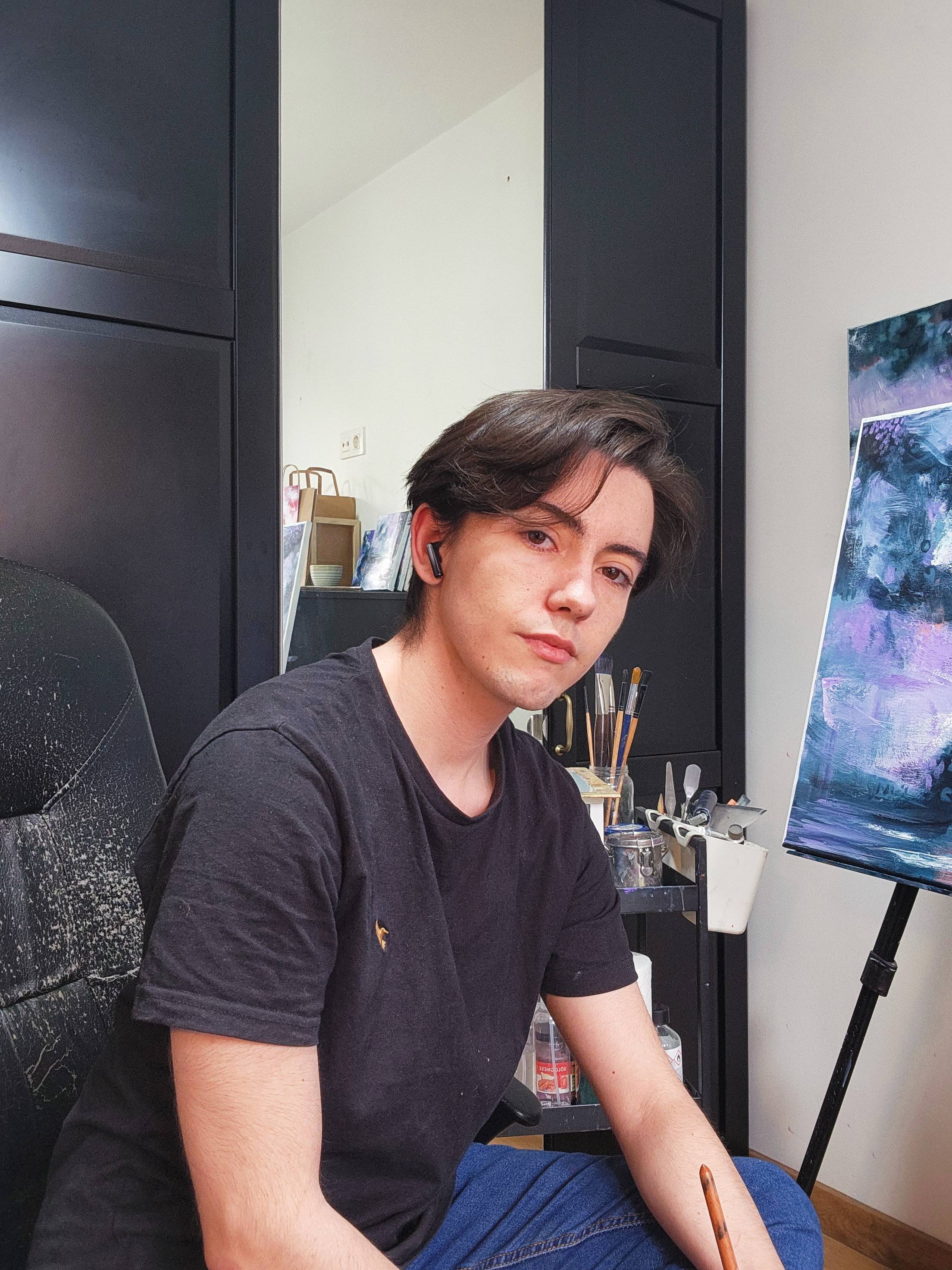 Joven hombre con cabello castaño oscuro, usando una camiseta negra, sentado en una silla de oficina en un estudio de arte, con una mesa de herramientas y un lienzo con pintura abstracta a su lado.