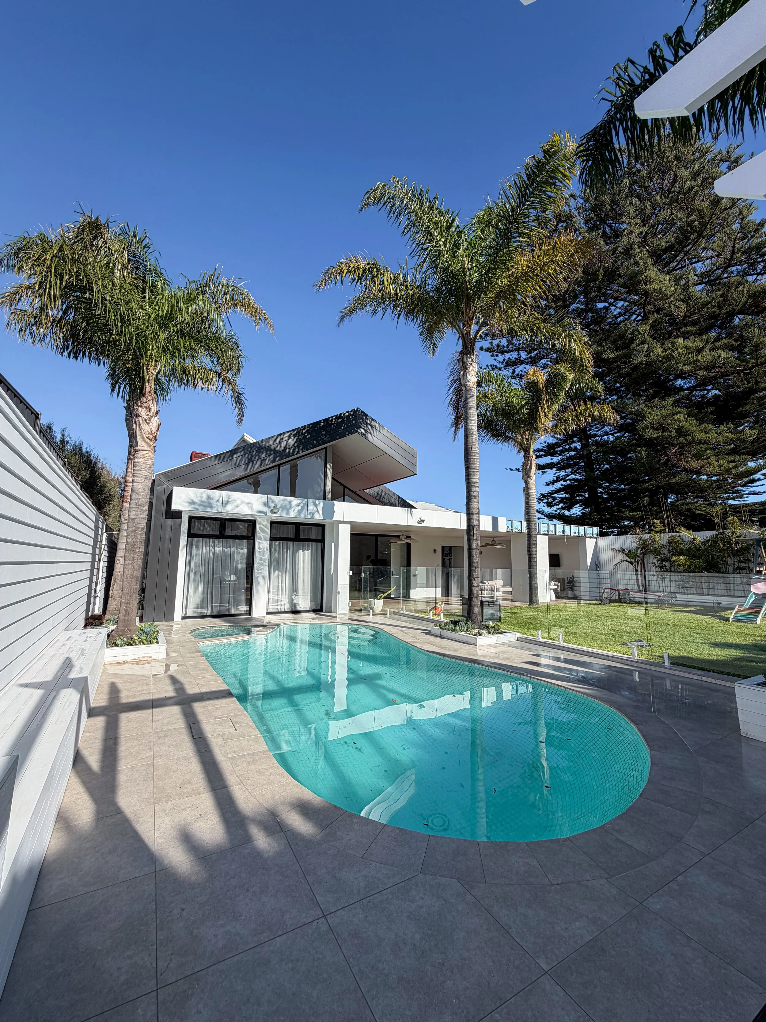 Modern house with a backyard swimming pool, tall palm trees, lush greenery, and a clear blue sky.