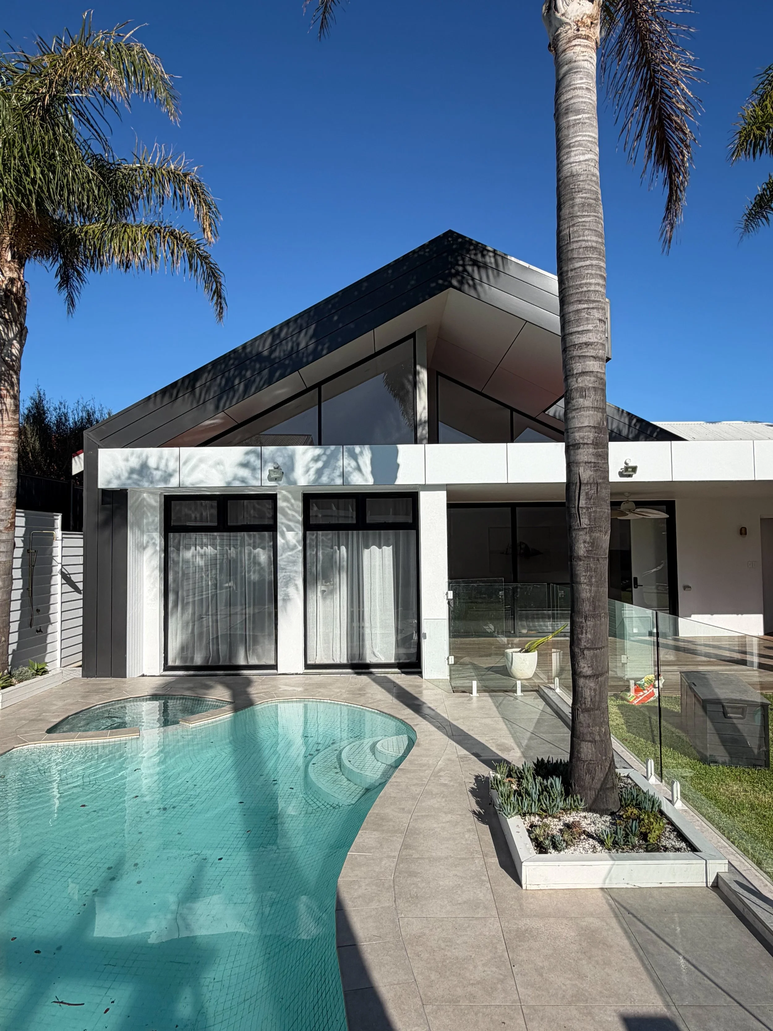 Modern house with large glass windows, a sloped roof, a small swimming pool in the backyard, and tall palm trees under a clear blue sky.