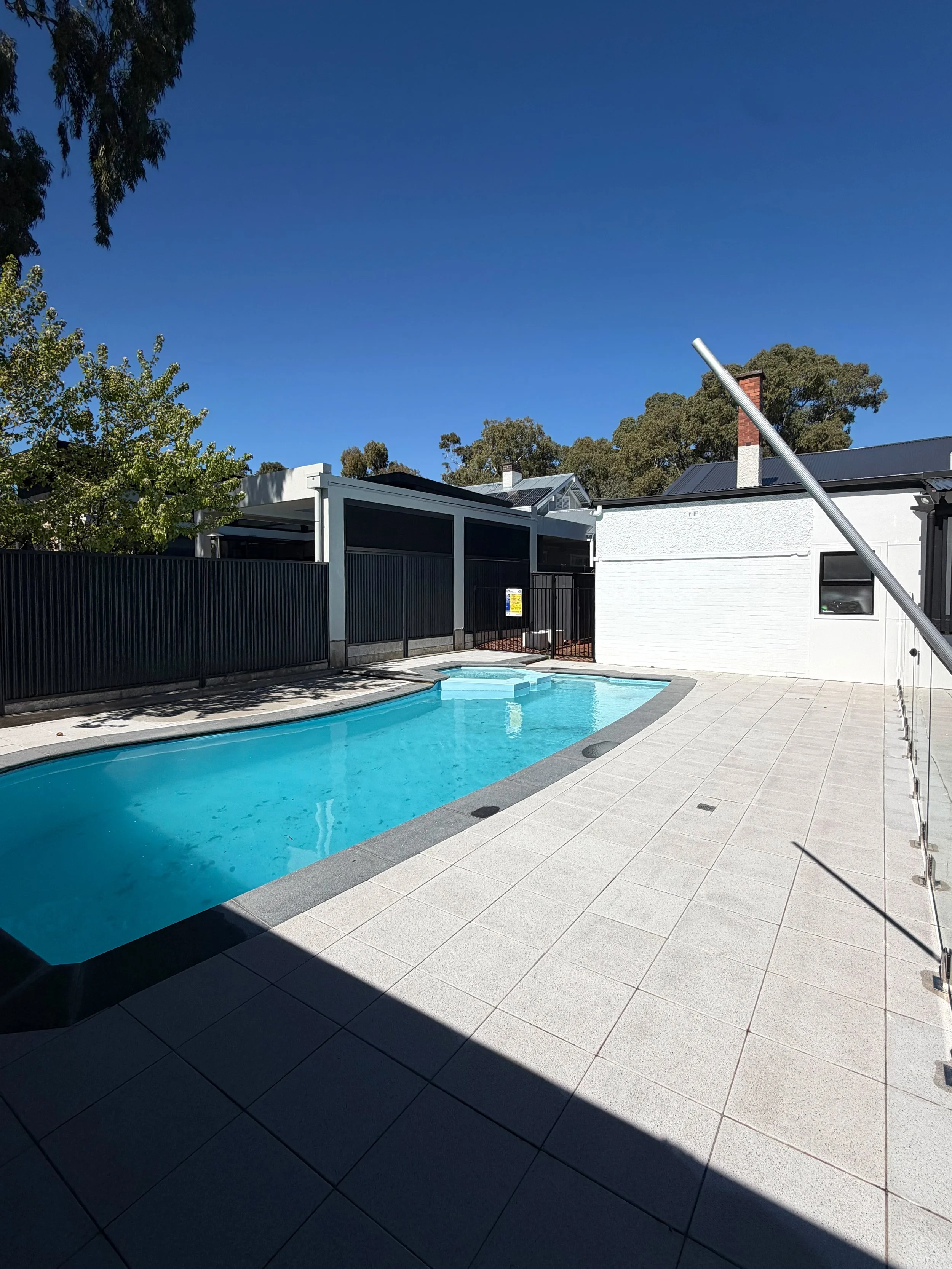 Residential backyard with a swimming pool, surrounded by a tiled patio, a black fence, and houses with greenery and trees in the background under a clear blue sky.