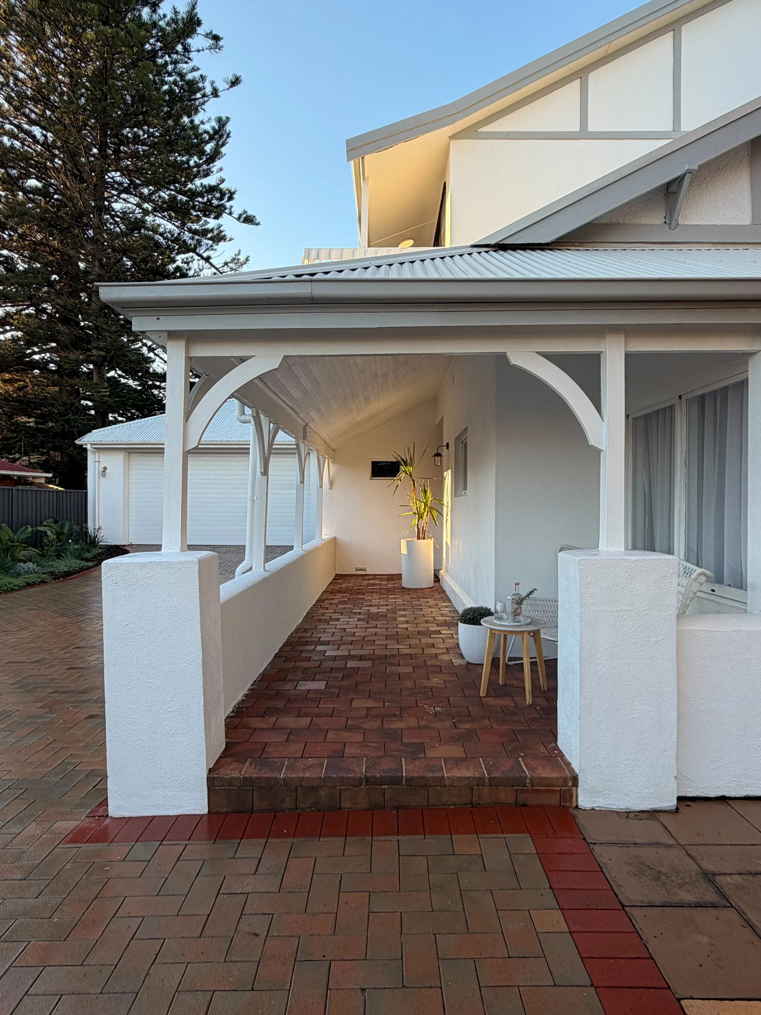 Exterior of a modern white house with a covered porch, brick pathway, potted plants, and a large tree in the background.